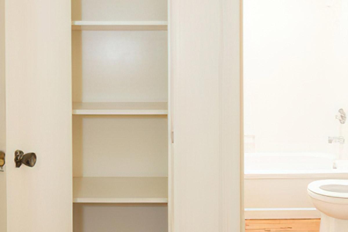 A white cabinet with open doors revealing empty shelves, situated next to a bathroom door and a toilet. The interior features light-colored walls and wooden flooring.