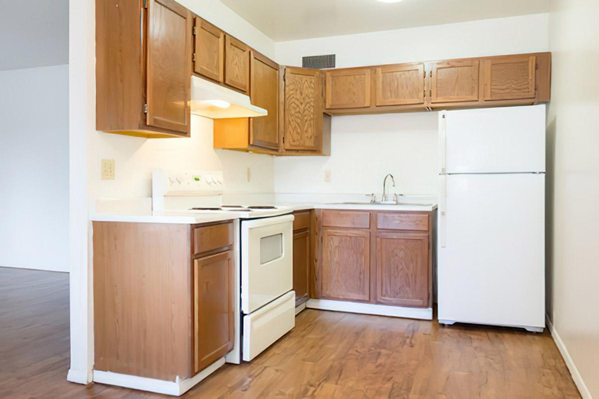 A small kitchen featuring wooden cabinets, a white stove, an oven, a sink, and a refrigerator. The countertops are light-colored, and the flooring is wood. The space is well-lit, with a minimalistic design and an open layout leading to another room.