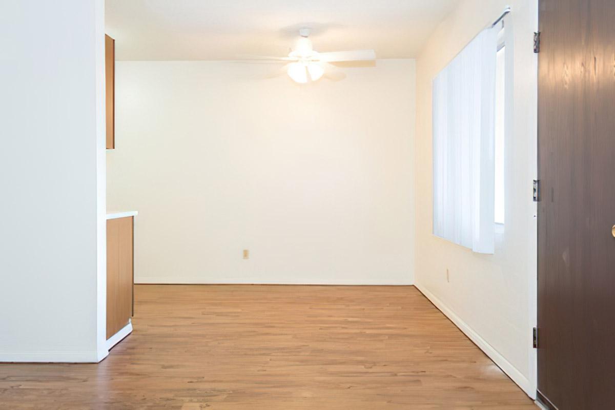 A spacious, empty room featuring light-colored walls and wooden flooring. A ceiling fan is visible in the center, and there are large windows with white shades allowing natural light to enter. There is a brown door on one side and a small kitchenette area partially visible to the left.