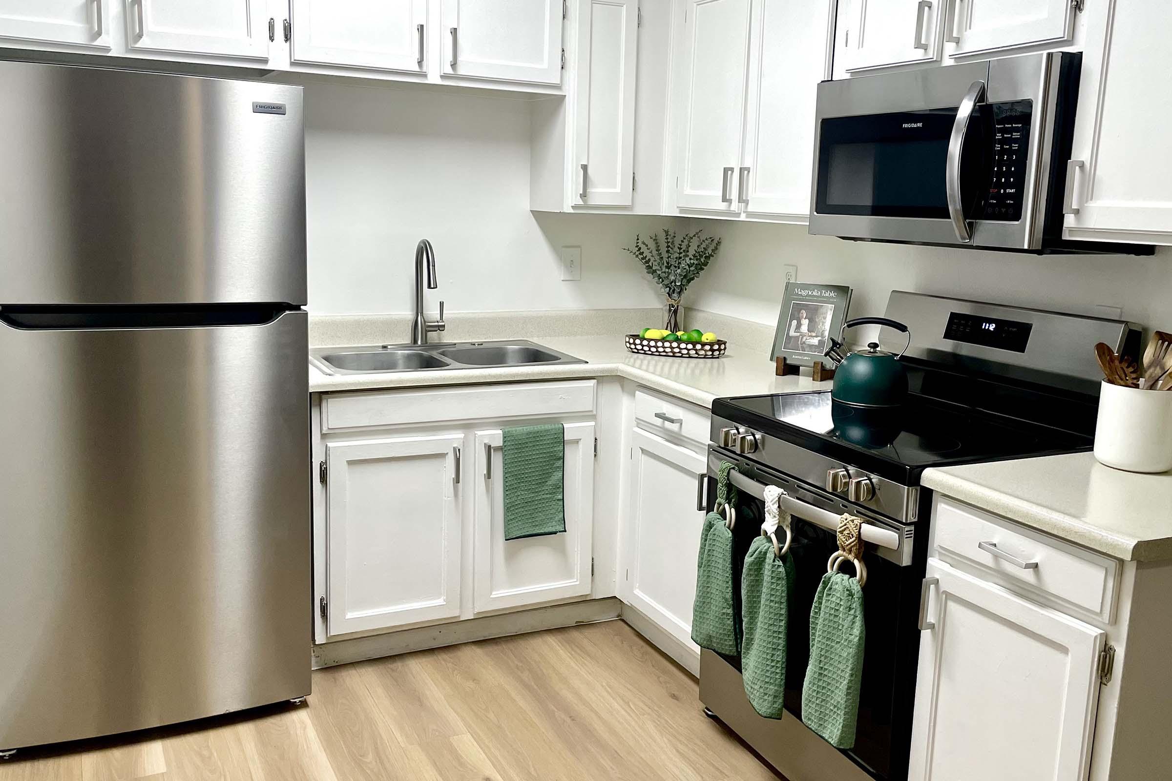 Modern kitchen featuring stainless steel fridge, black stove, and microwave. Light wood flooring complements white cabinets and countertops. A mint green kettle sits on the stove, while green dish towels hang from the oven. A decorative bowl with fruit and a small plant add a touch of color to the countertop.