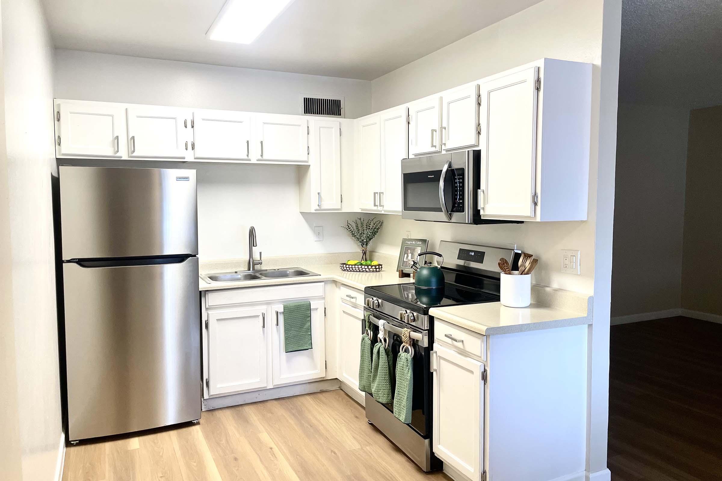 Modern kitchen featuring white cabinetry, stainless steel appliances, a sink, and a stove. Green dish towels hang on the oven. There's a decorative plant on the countertop and a bowl of fruit. Light wood flooring extends into an adjacent room with beige walls. A bright and inviting space with ample natural light.