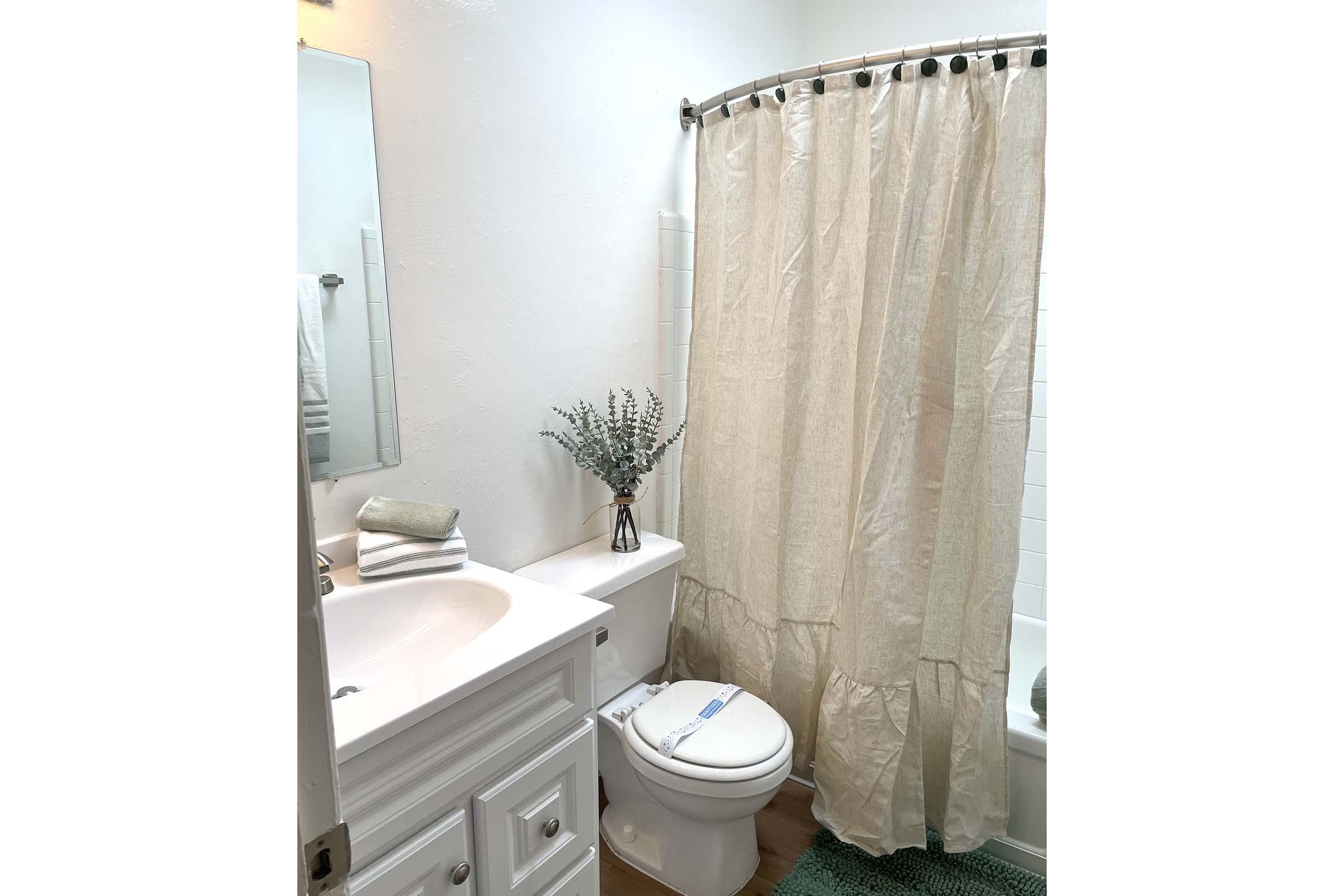 A bright, clean bathroom featuring a white shower curtain, a small vase with greenery on the sink, folded towels, and a light-colored rug. The bathroom has a neutral color scheme with a bathtub, toilet, and a wall-mounted mirror above the sink.