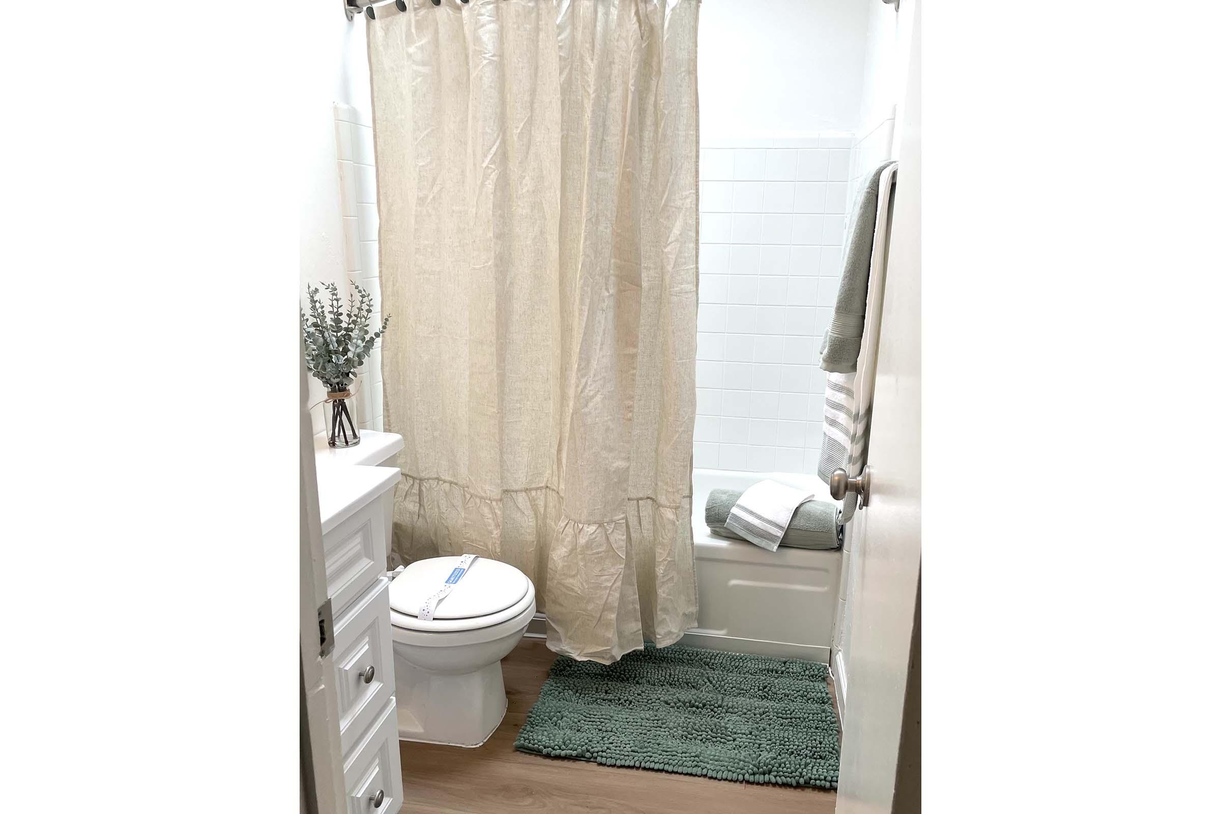 A clean bathroom featuring a white shower curtain, a toilet, and a light green bath mat. There are fluffy towels on a rack, a small plant in a vase on the counter, and white tiles on the walls. The overall decor is simple and inviting, with natural light illuminating the space.