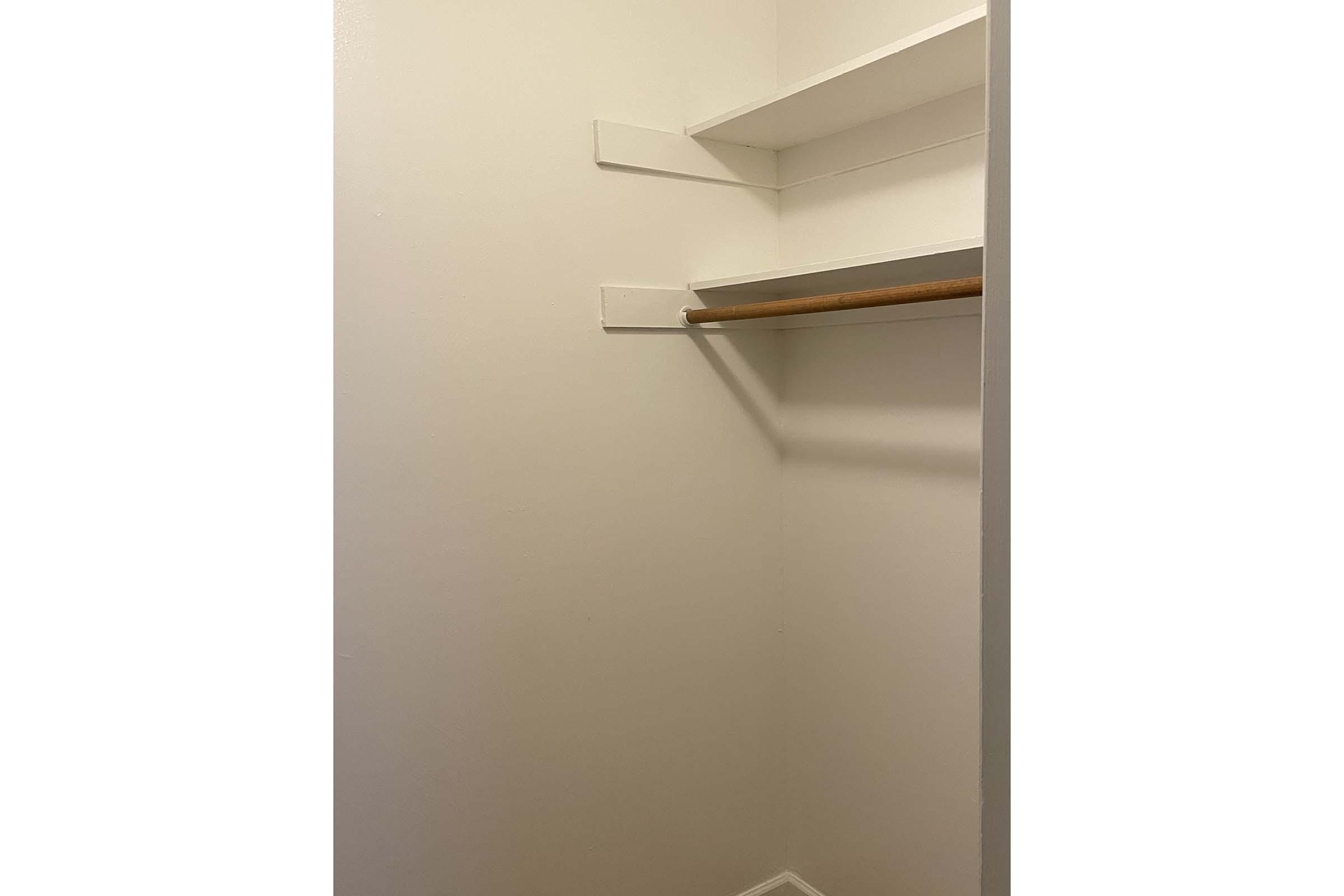 Interior view of a minimalist closet featuring a light-colored wall, a wooden clothing rod, and two empty shelves. The space is uncluttered and designed for storage, with a clean, simple aesthetic. The floor remains bare, emphasizing the emptiness of the closet.