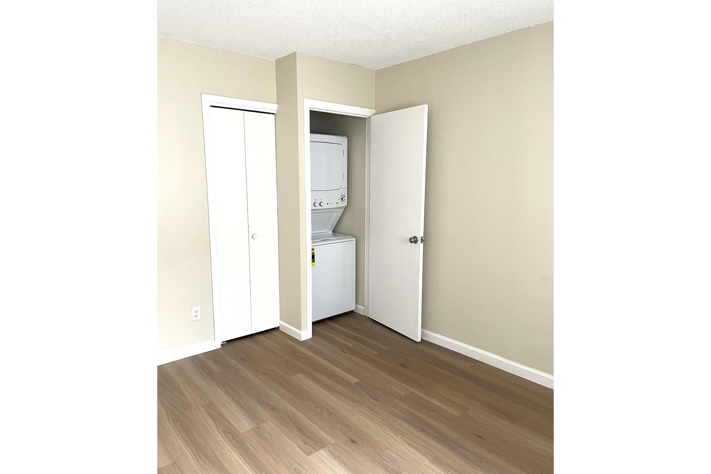 A small interior space with light-colored walls and hardwood flooring. There is a closet on the left and an open door leading to a laundry area with a stacked washer and dryer. The room appears well-lit and empty, suitable for various uses.