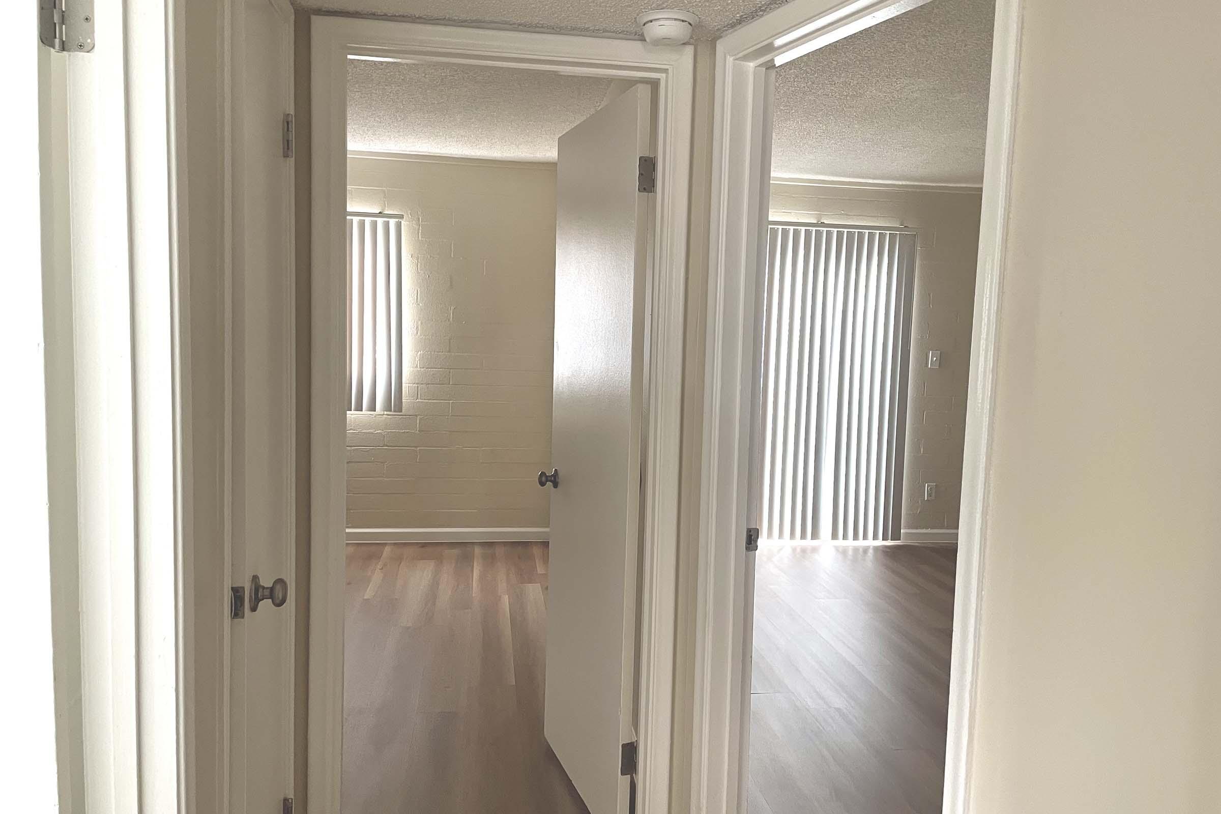 A view of an interior hallway with two open doors leading to different rooms. The walls are painted light colors, and the floor is wooden. Natural light filters through vertical blinds on the windows. One room appears empty while the other has a neutral atmosphere, creating a spacious feel.