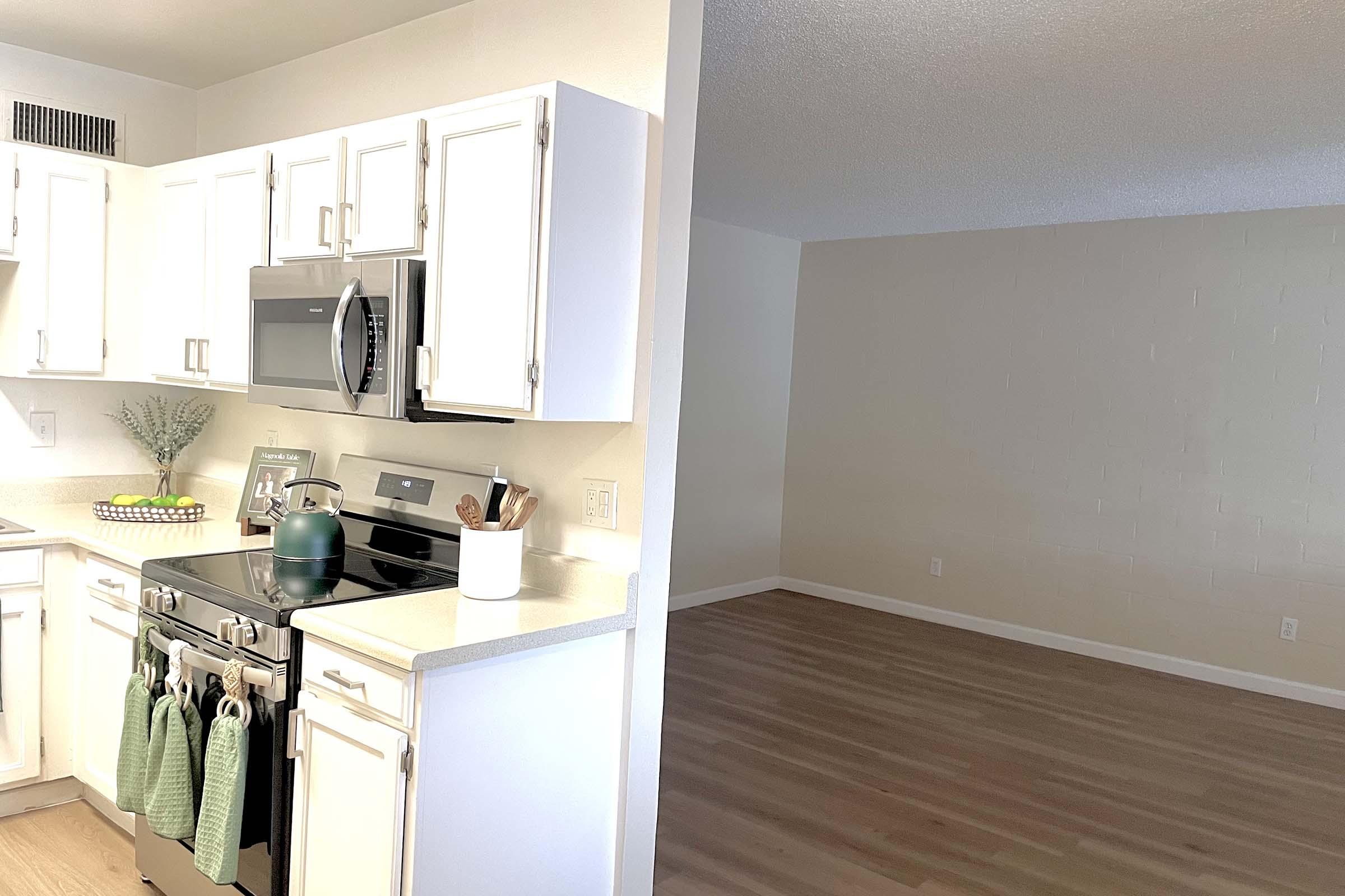 Modern kitchen with white cabinetry, stainless steel appliances, and a stove. A green kettle sits on the counter. The adjacent space is an empty room with light-colored walls and wooden flooring, providing a bright and open atmosphere.