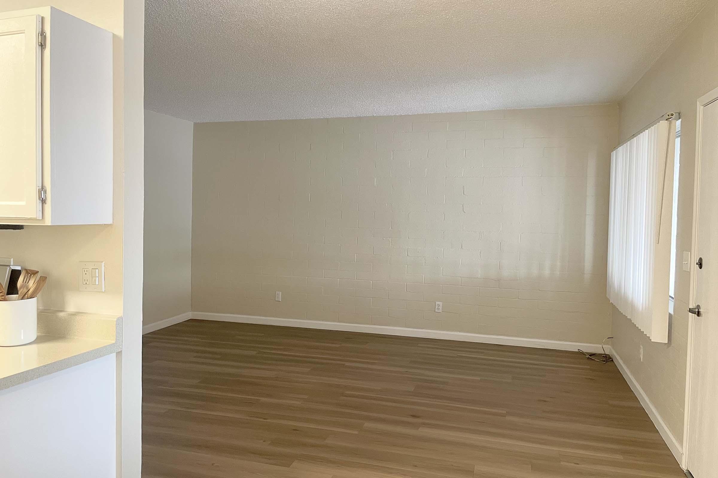 A spacious, empty room with light-colored walls and wooden flooring. The room features a large window with white curtains that lets in natural light. There is a doorway visible on the right, leading outside. The overall atmosphere is bright and inviting, ideal for personalizing with furniture.