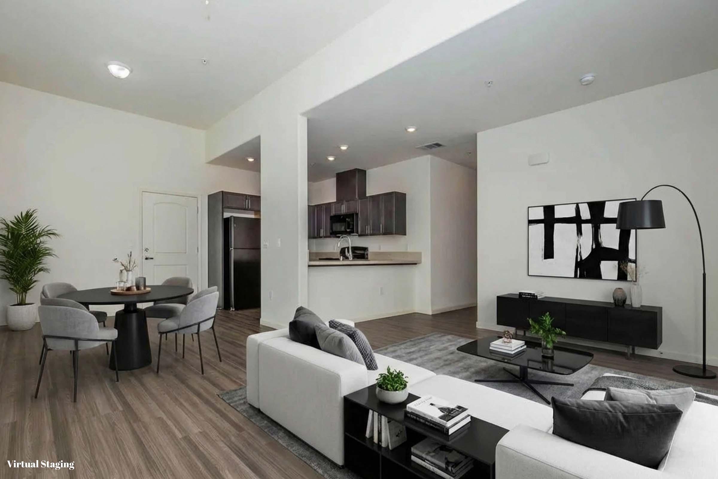 A modern living room and kitchen area featuring a light-colored sofa, a round dining table with chairs, and a large flat-screen TV. The space has hardwood flooring, minimalistic decor, and large windows providing natural light. Contemporary art is displayed on the walls, creating an inviting atmosphere.