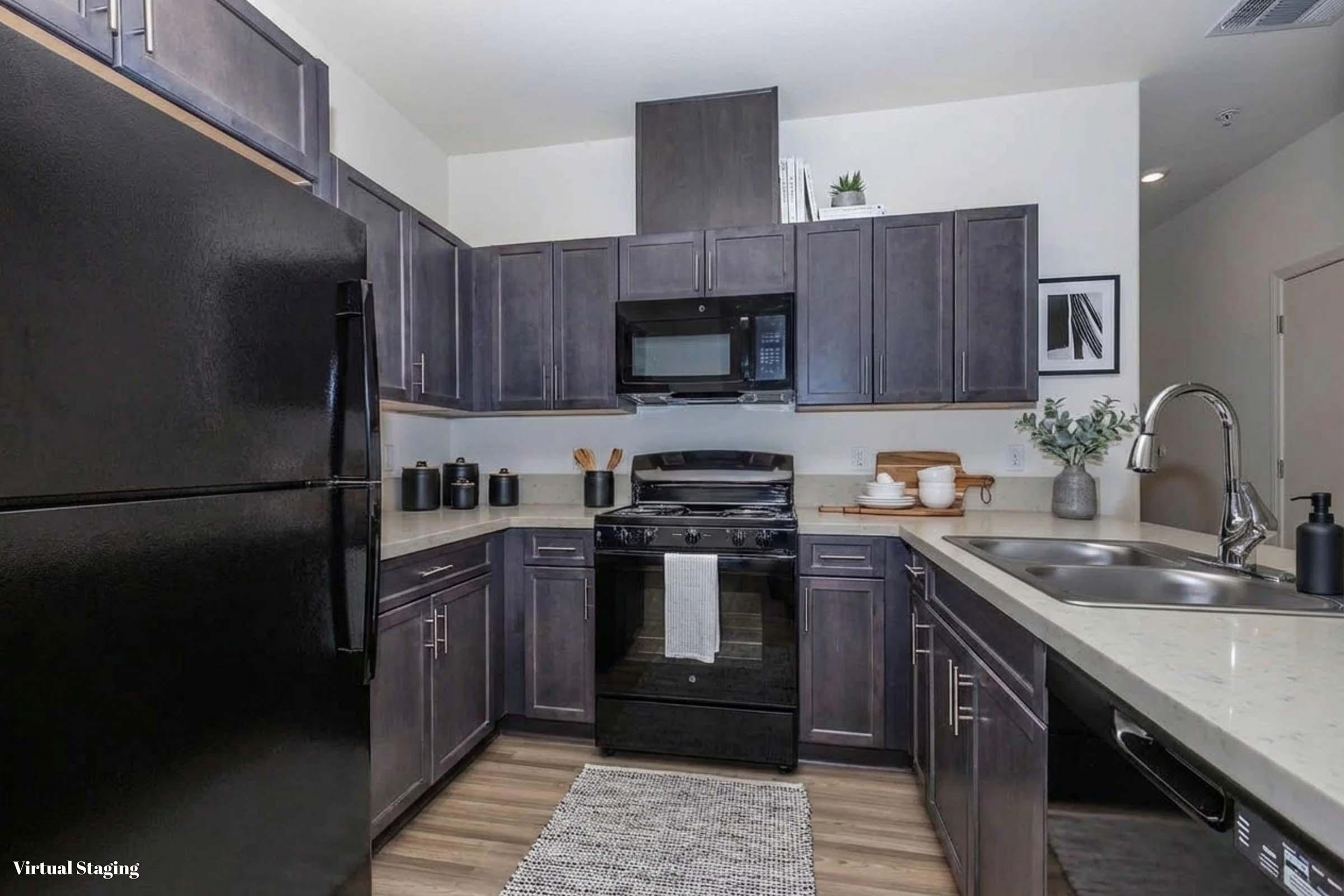 Modern kitchen with dark wooden cabinets, stainless steel appliances, and a countertop. A black refrigerator, gas stove, and microwave are visible. The decor includes greenery and dishware arranged neatly. A light-colored rug lies on the floor, and the space is well-lit with a clean, contemporary design.