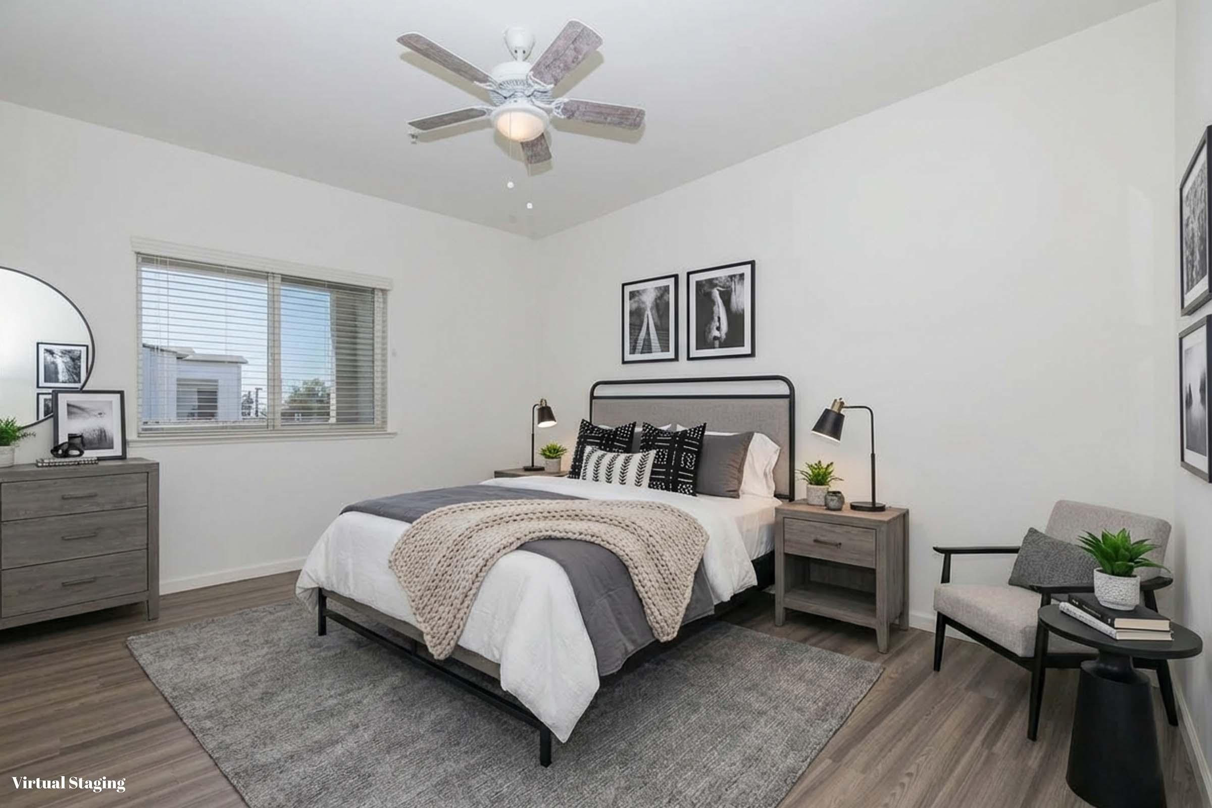 A modern bedroom featuring a bed with a gray and white blanket, two decorative pillows, and a nightstand. A large round mirror is mounted on the wall, along with framed black-and-white photographs. There is a small armchair and potted plants. A ceiling fan is visible above. The walls are light-colored, enhancing the spacious feel.