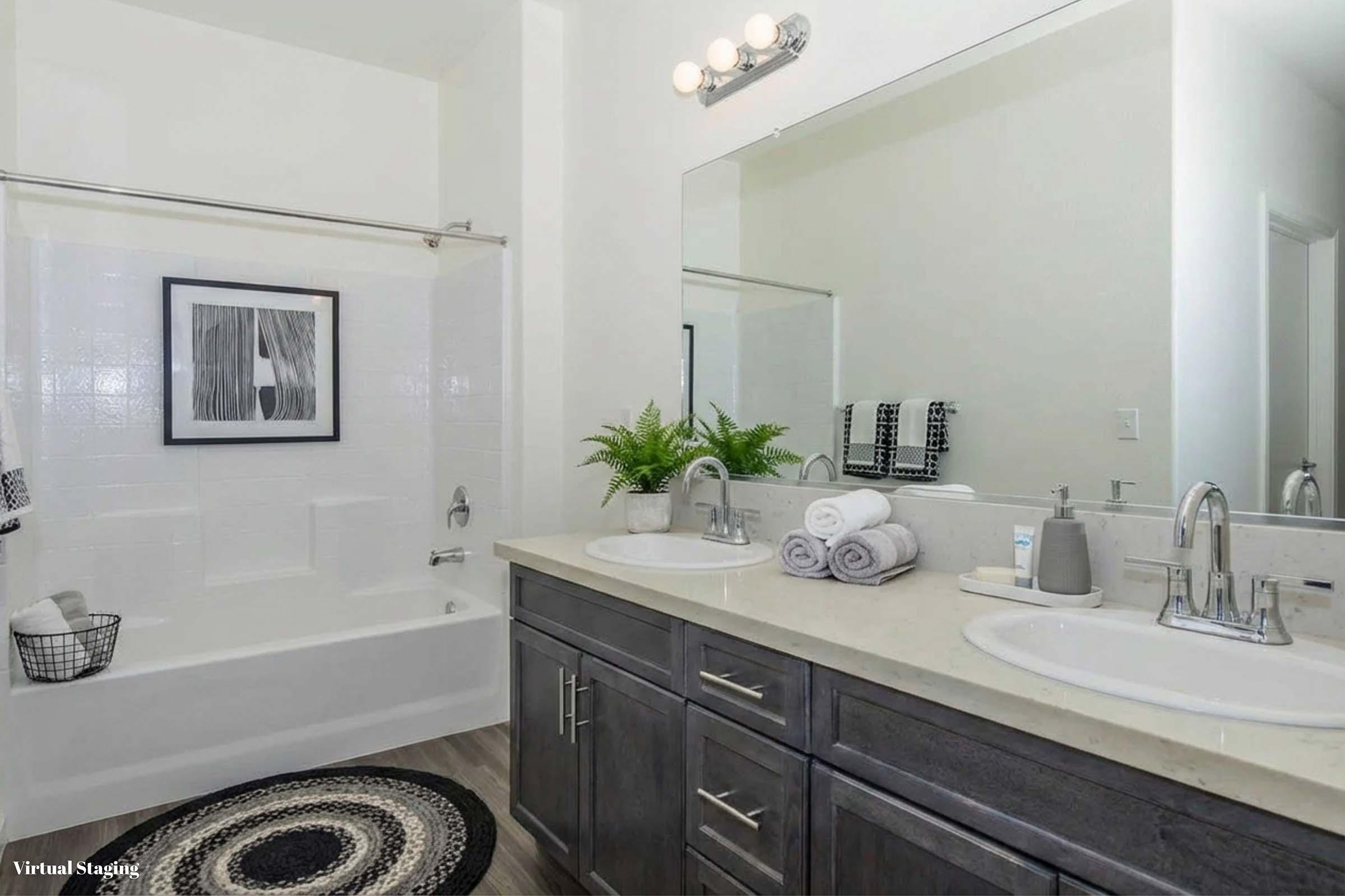 A modern bathroom featuring a double vanity with silver fixtures, a large mirror, and towels neatly arranged. The bathtub is accompanied by a black and white artwork. A potted plant adds a touch of greenery, while a round rug lies on the floor, enhancing the contemporary style of the space.