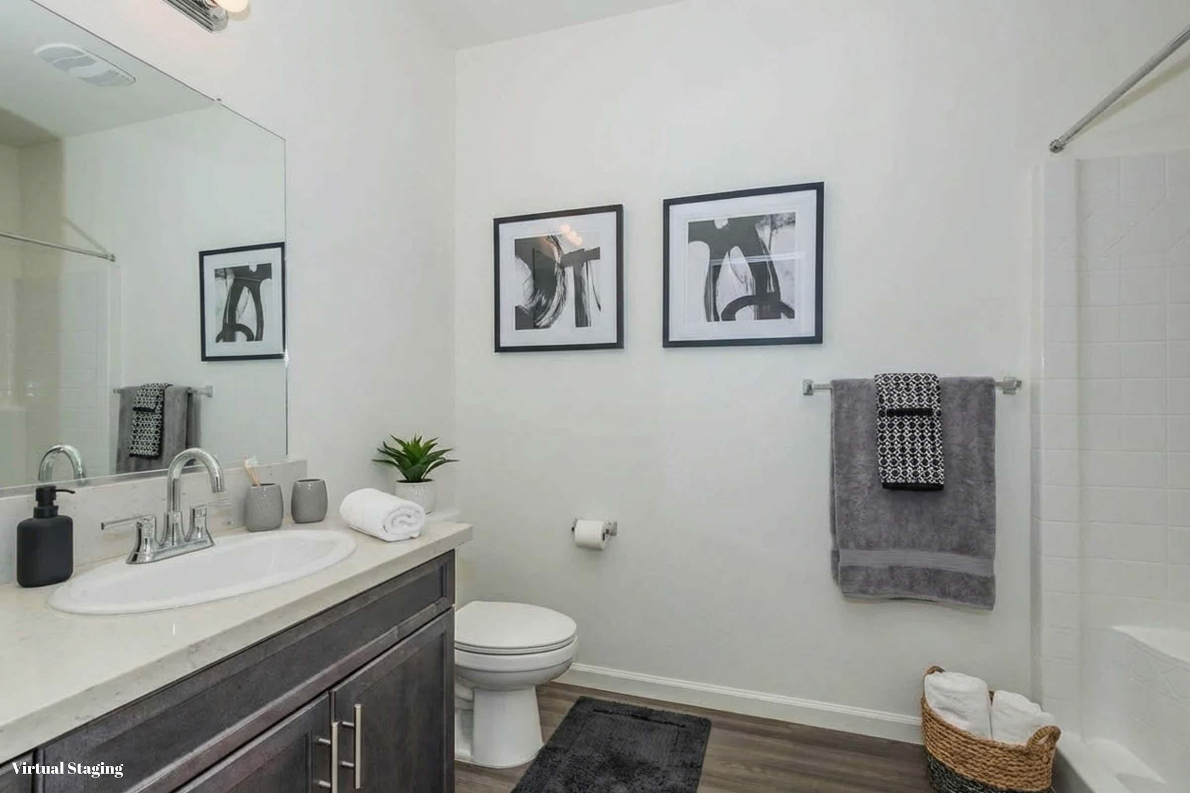 A modern bathroom featuring a white wall, dark wood cabinetry, and a white toilet. Two abstract framed prints hang above the sink, which is equipped with a faucet and soap dispenser. A plush gray towel is hung on a rack, while an additional towel and a small plant are placed nearby.