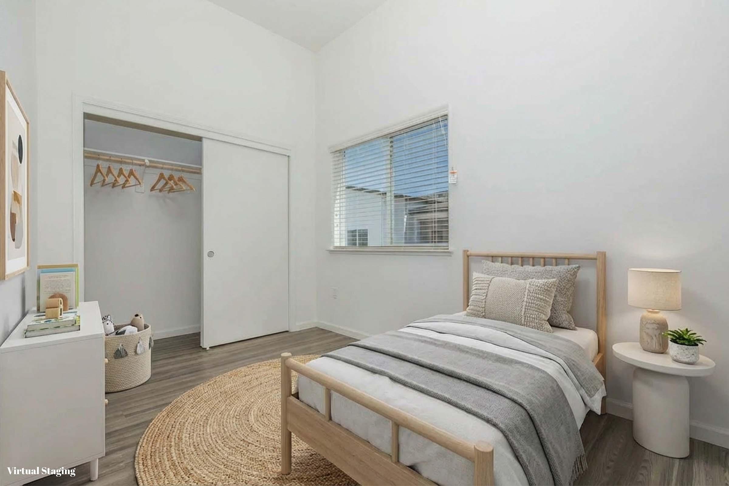A cozy bedroom featuring light-colored walls and wooden flooring. The room includes a single bed with gray and white bedding, a round jute rug, a small table beside the bed, and a closet with sliding doors. A window lets in natural light, highlighting the minimalist decor.