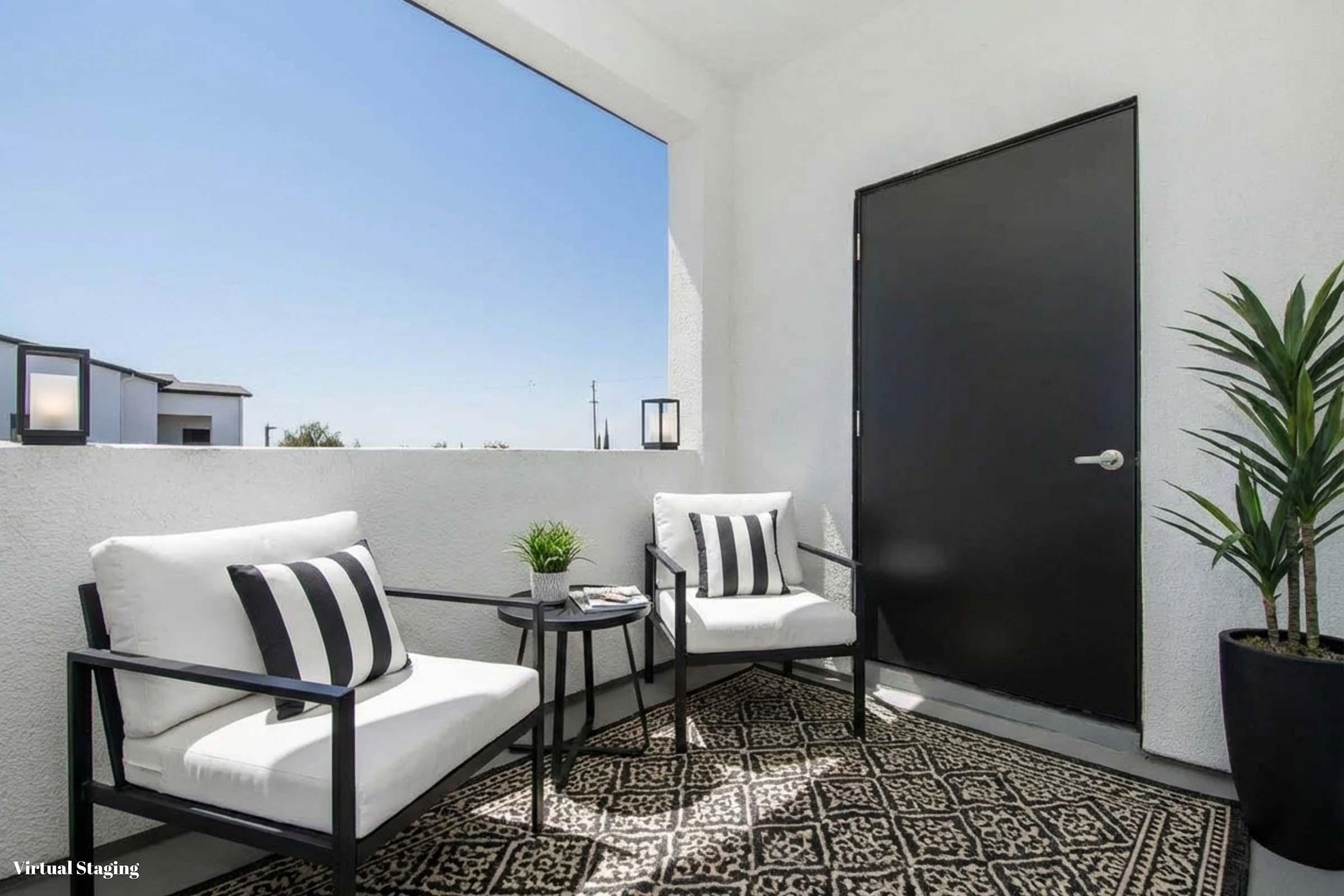 A small balcony featuring two black and white striped chairs beside a round table. A potted plant is in one corner, and a large black door is visible, set against a bright blue sky. The floor is adorned with a decorative rug, creating a cozy outdoor space.