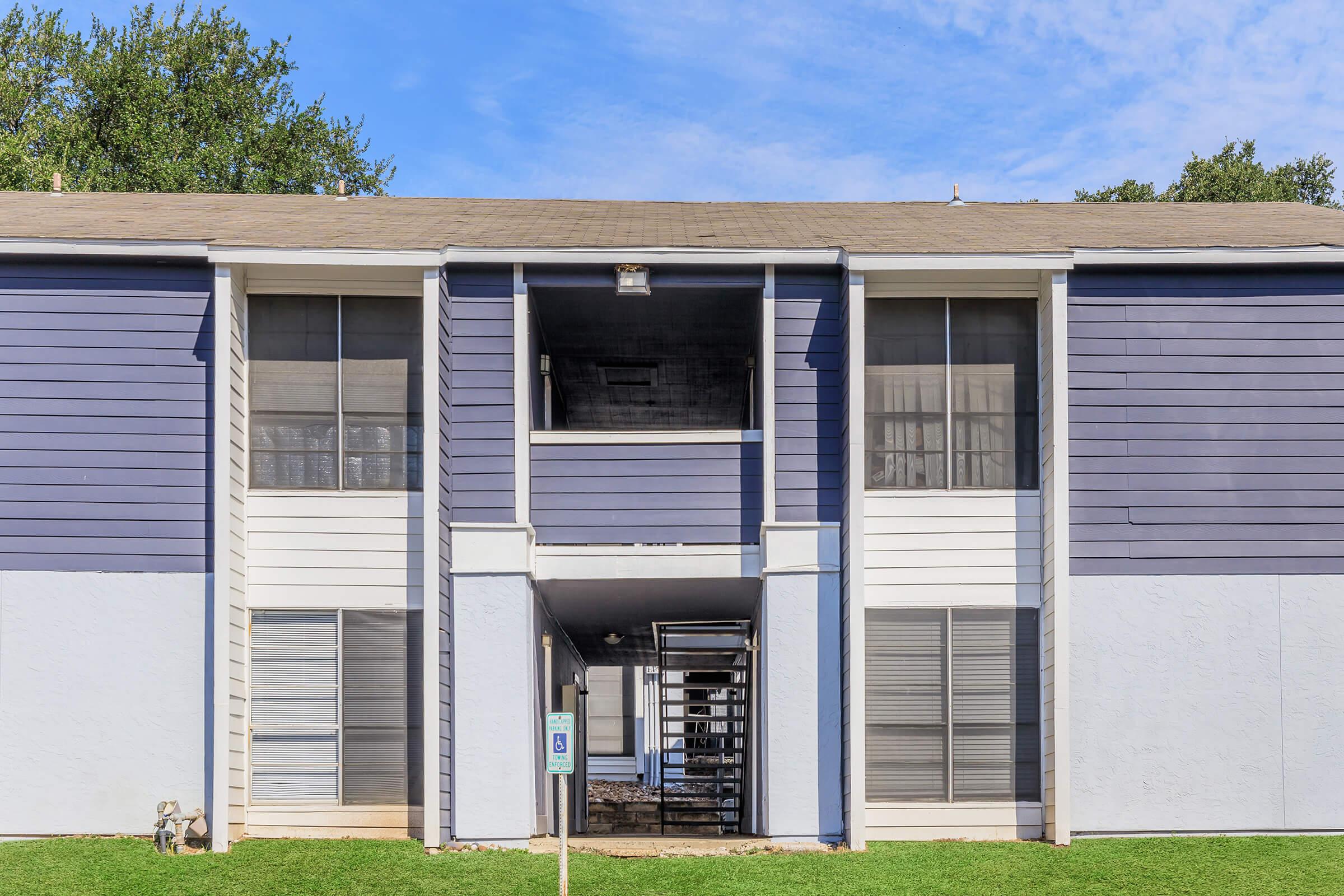 Exterior view of an apartment building featuring two stories, with blue and white siding. The entrance is framed by a covered balcony, flanked by large windows. Grass is visible in the foreground, and the sky is bright with scattered clouds. A parking sign is located nearby.