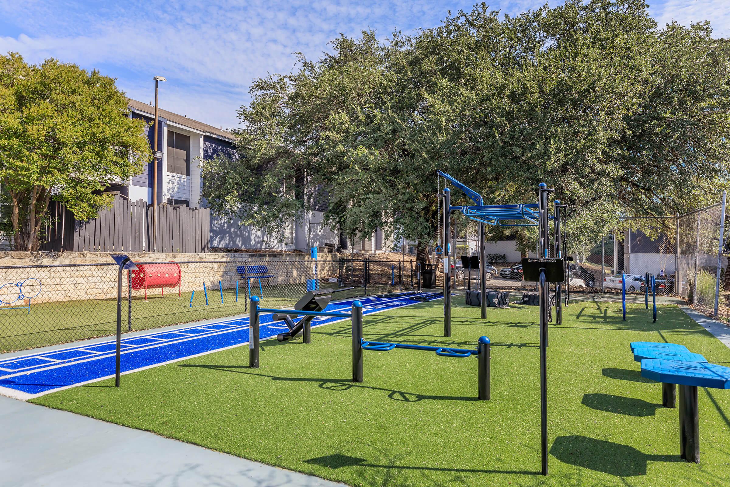 A vibrant playground featuring blue and green turf, equipped with a swinging area, climbing structures, and exercise equipment. Surrounded by trees and benches, it offers a spacious setting for outdoor activities, with a residential building in the background and a clear blue sky above.