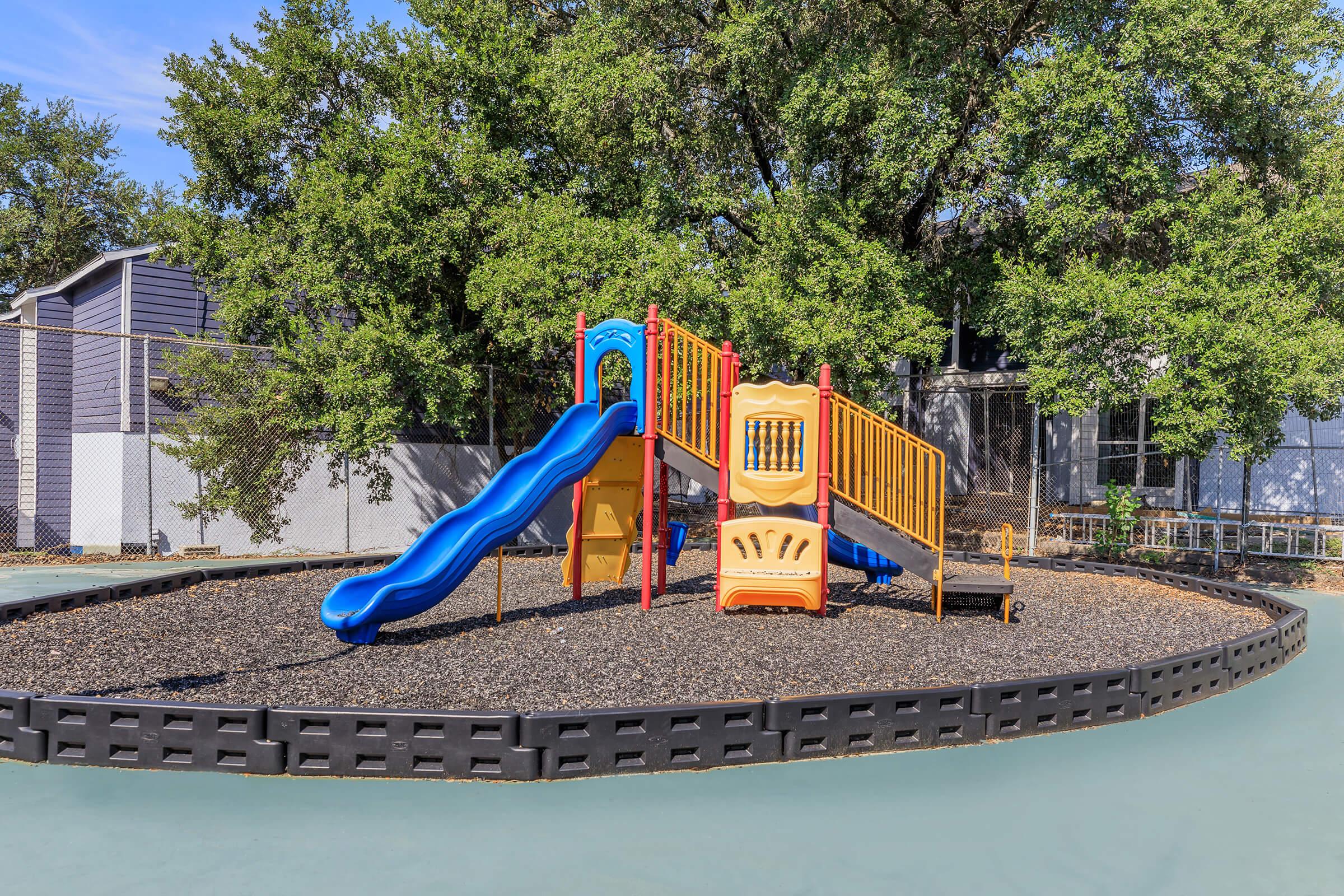 A colorful playground featuring a yellow and orange play structure with slides, surrounded by a soft black ground cover. Lush green trees provide shade in the background, with a fence enclosing the area. The setting is bright and inviting for children to play.