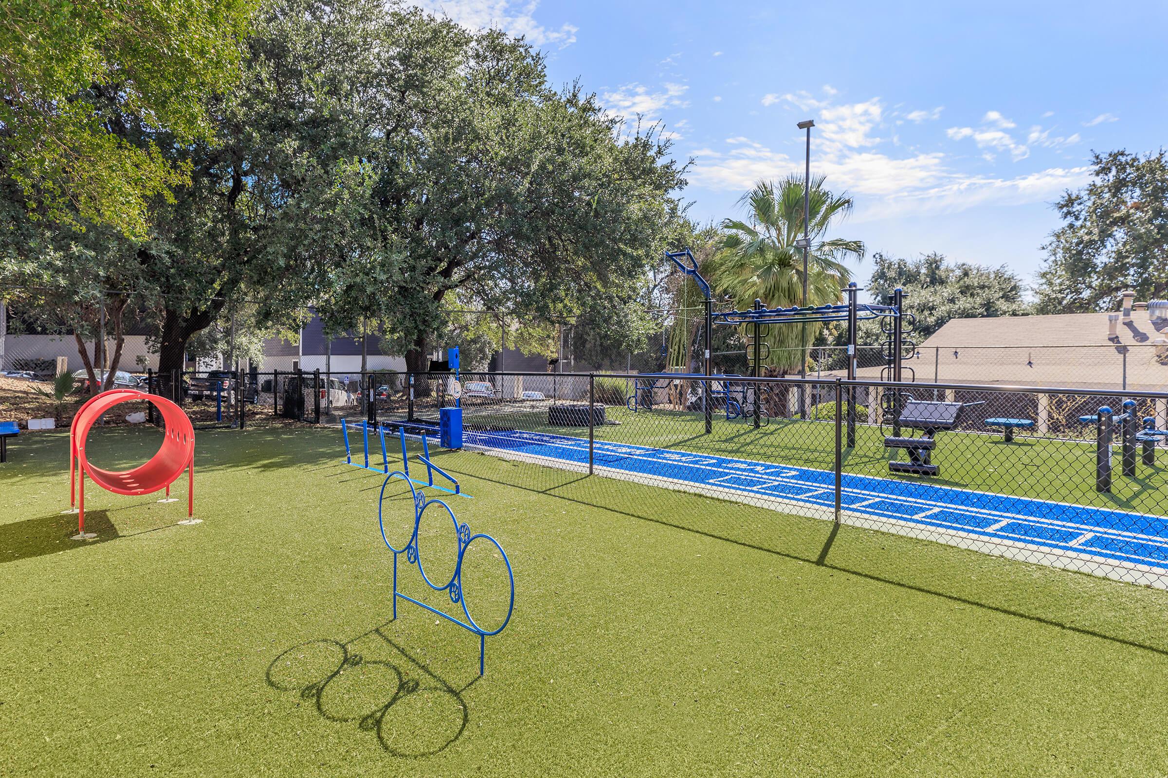 Playground area featuring a blue climbing structure, circular tunnel, and agility equipment on a green synthetic turf surface. Surrounded by trees and a fence, with benches and exercise stations visible in the background under a clear blue sky.