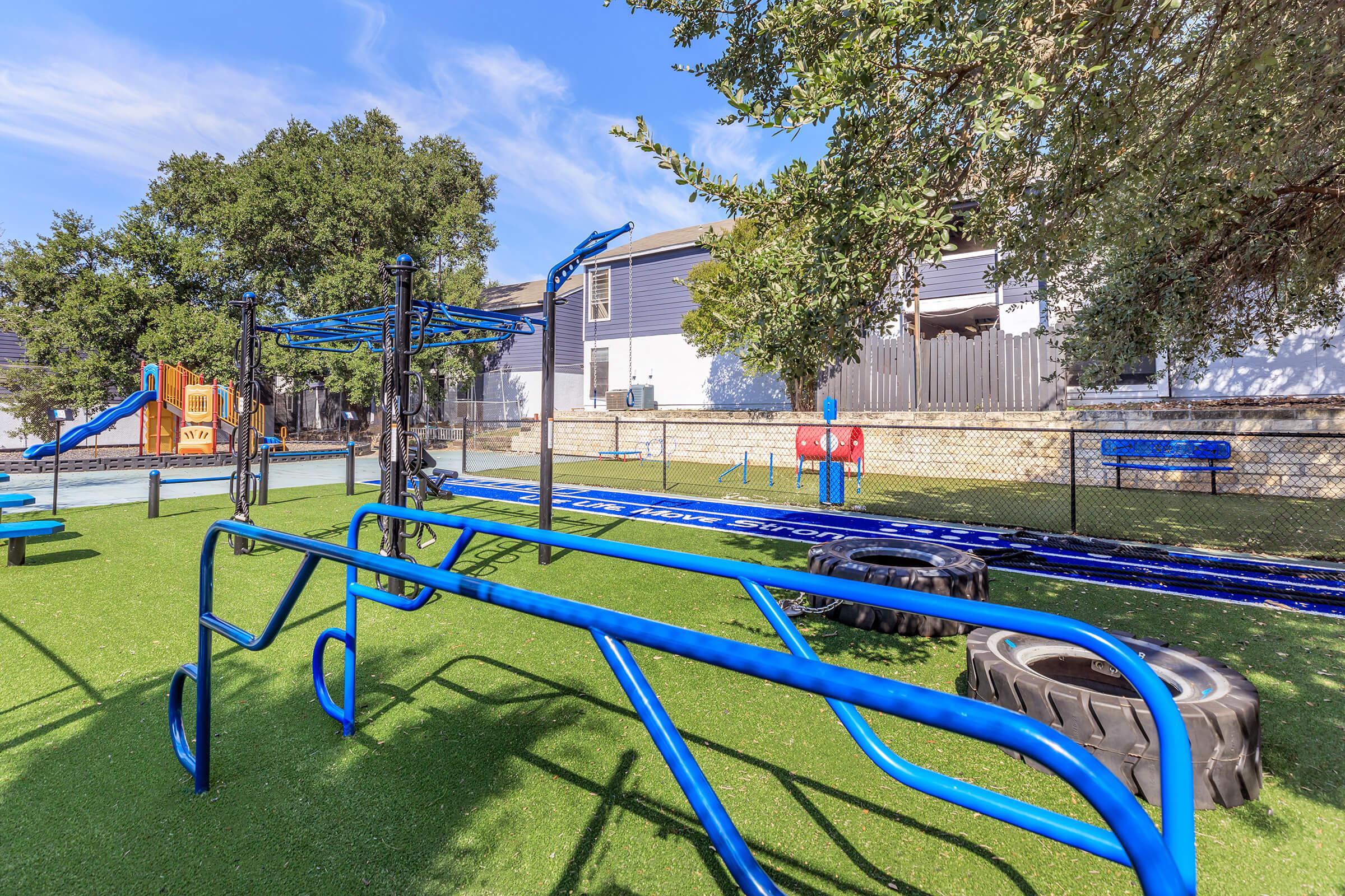 A colorful playground featuring blue fitness equipment, a climbing structure, and tires for exercise, surrounded by green artificial turf. The area is enclosed with a fence and is set against a backdrop of residential buildings and blue skies.