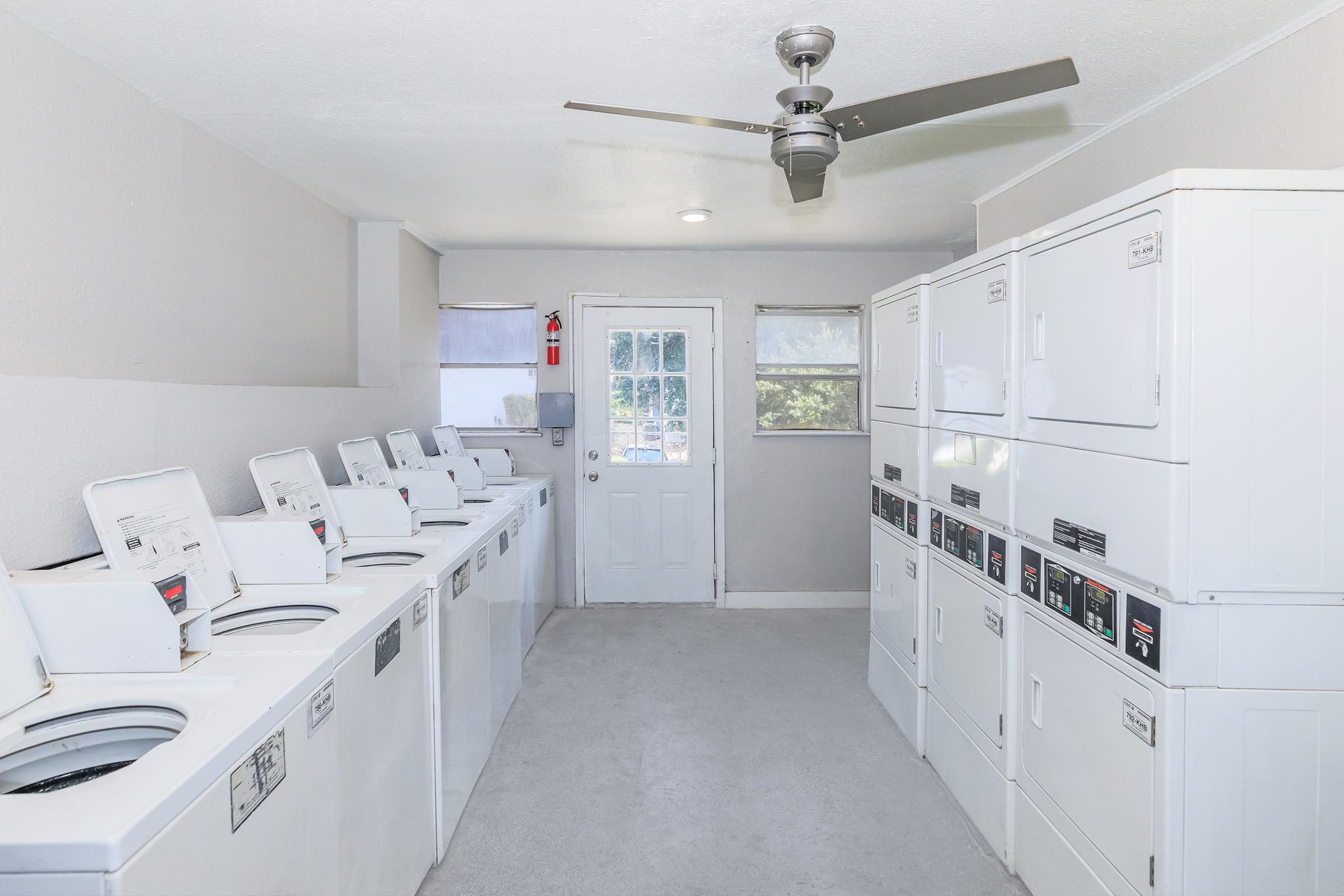 A bright, clean laundry room featuring multiple white washing machines and dryers lined against the walls. A door leads to the outside, and there's a ceiling fan overhead. Windows allow natural light to fill the space, creating an inviting atmosphere for doing laundry.