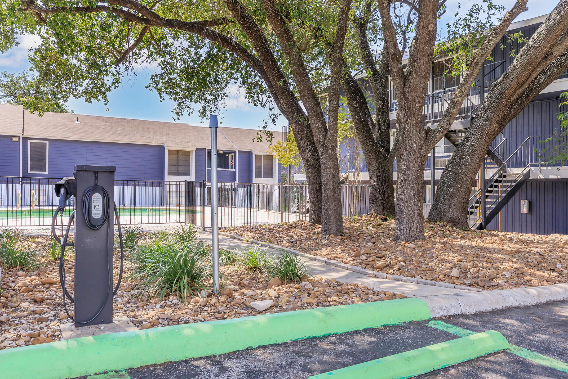 A parking area featuring a black electric vehicle charging station surrounded by green landscaping. Large trees provide shade, and a fenced area with a multi-story building is visible in the background. The pavement is marked with green lines indicating parking spots.
