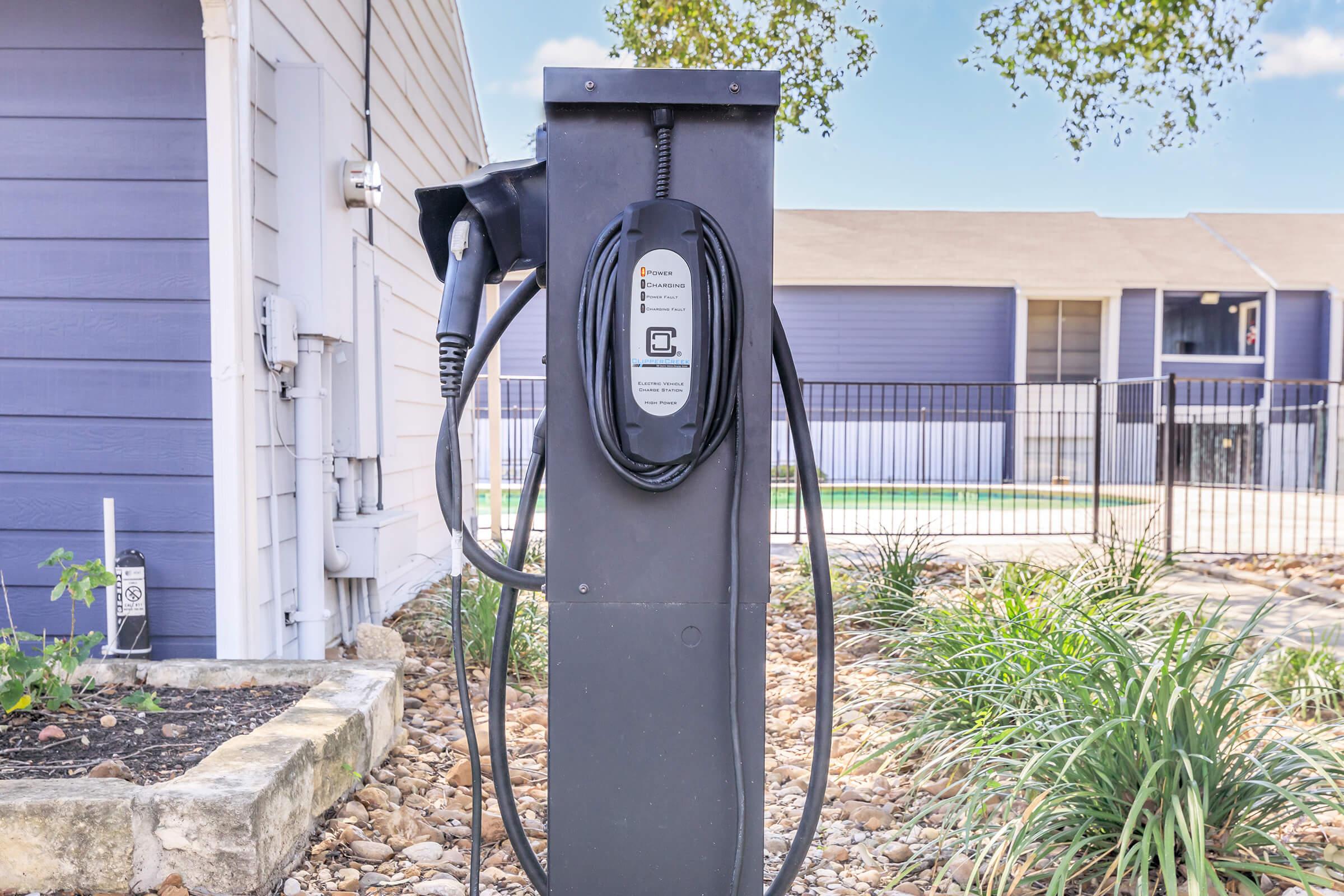 An electric vehicle charging station with a charging cable attached, set against a backdrop of a modern residential building and green landscaping. The station features a display panel for user instructions and is surrounded by decorative stones and plants.