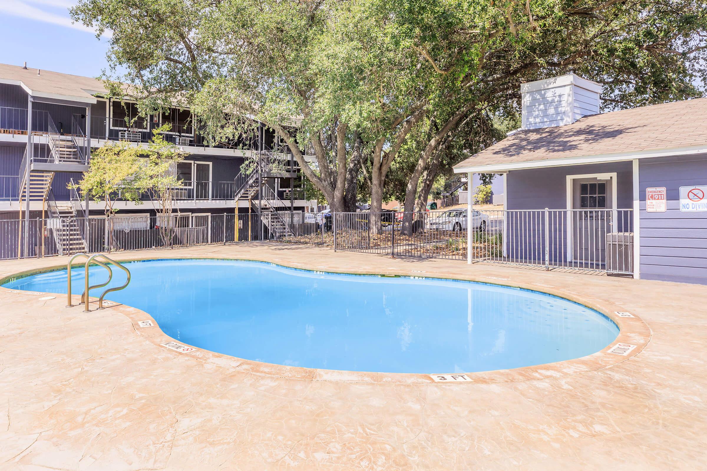 A clear blue swimming pool surrounded by a concrete deck and green trees. In the background, there are multi-story buildings with balconies. A small poolside building is visible, and there are no people in the scene. The area is well-lit and appears inviting.