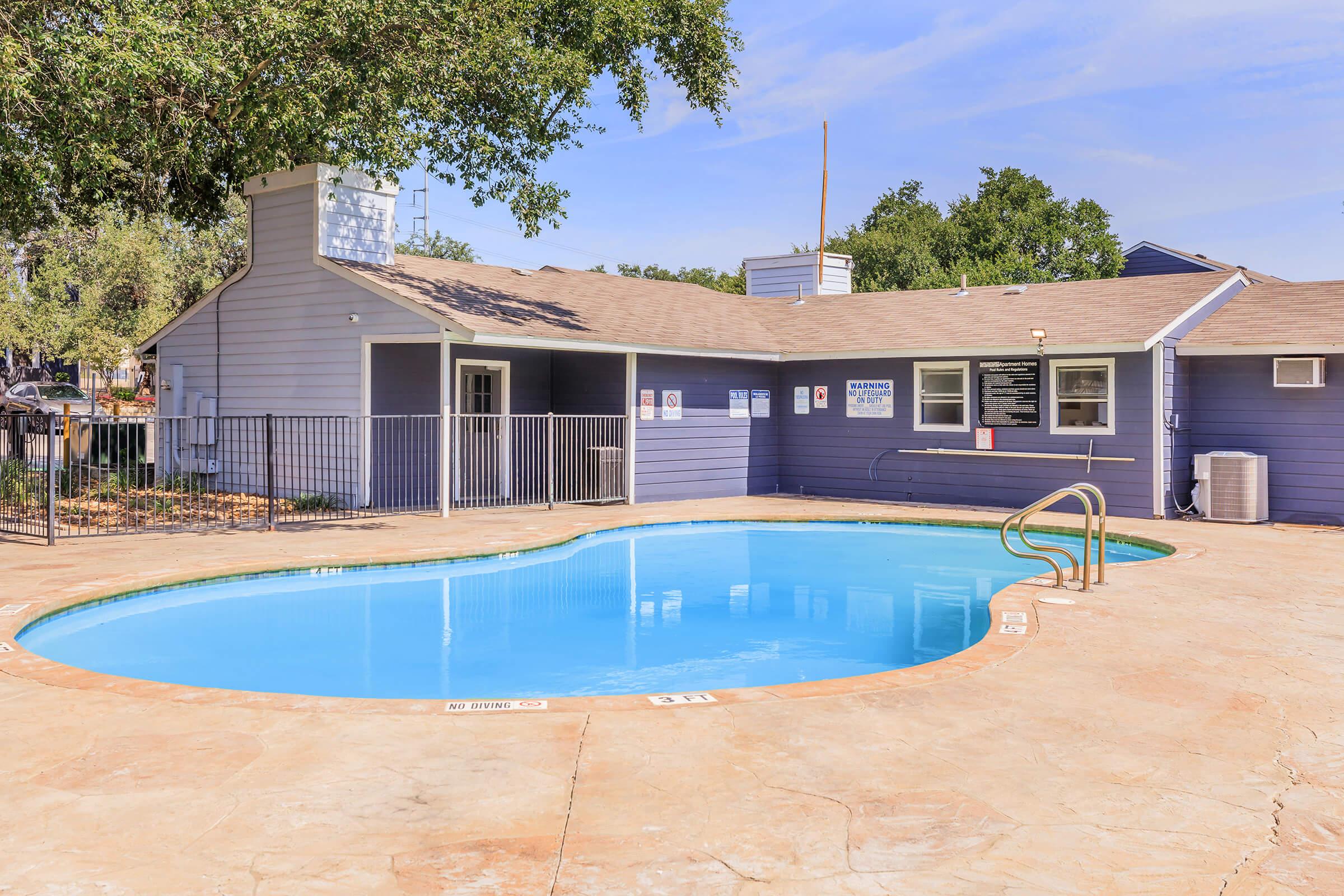 A swimming pool with a curved shape, surrounded by a concrete deck. In the background, a single-story building with a blue exterior and several windows can be seen, along with trees and a clear blue sky. A railing encloses the pool area, providing a secure space for relaxation and leisure.
