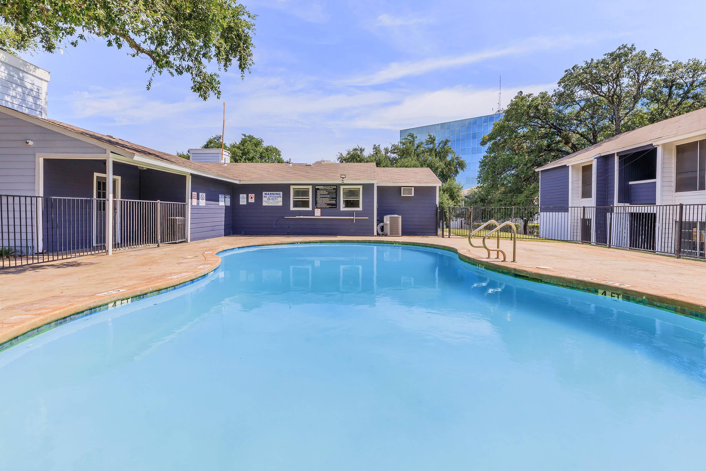 A clear blue swimming pool surrounded by a wooden deck. There are lounge chairs and a building with a sign nearby. Trees are visible in the background, along with a large glass building. The sky is partially cloudy, creating a bright and inviting atmosphere.