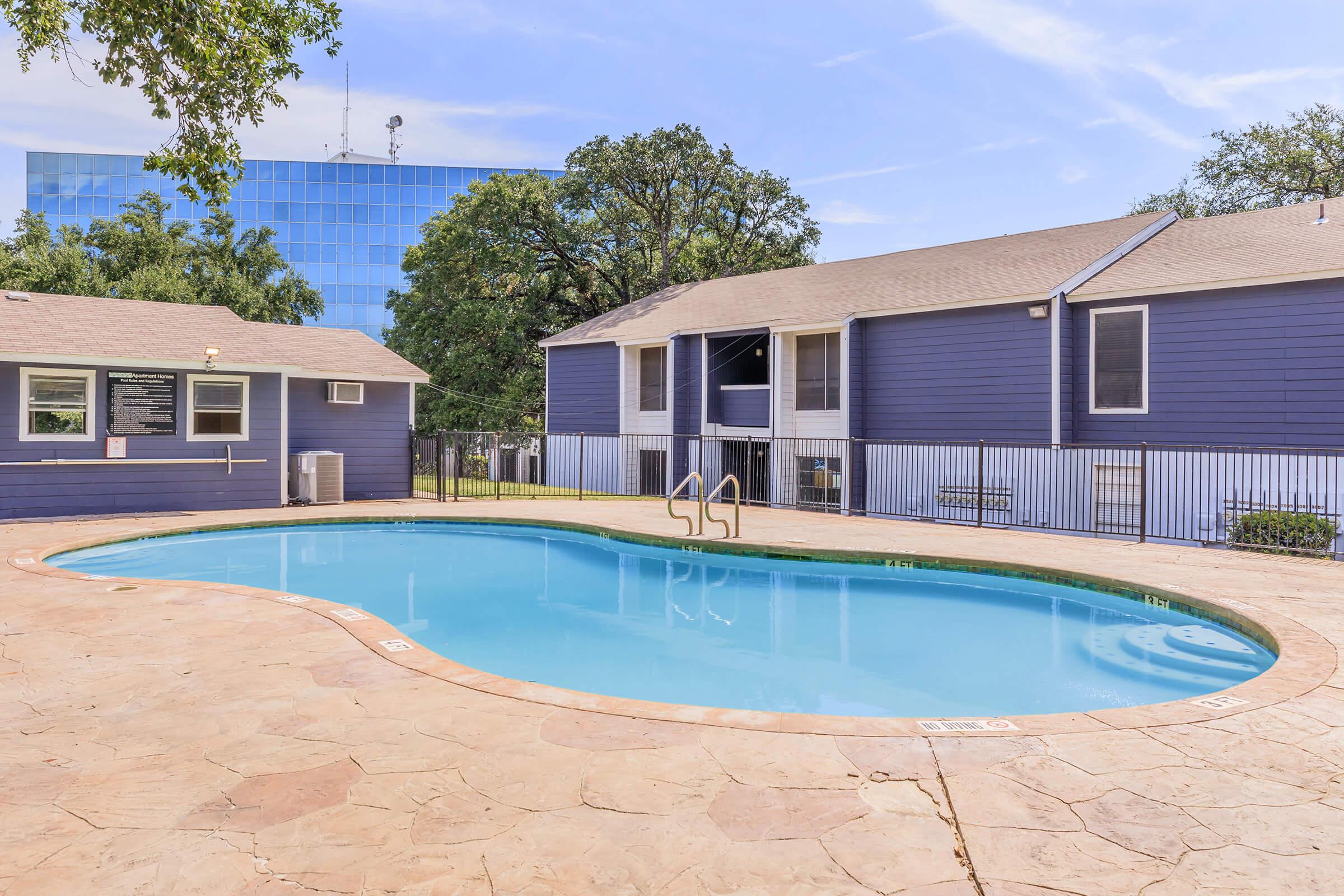 A serene outdoor pool surrounded by a stone patio, with a nearby blue two-story building and green trees. The pool is fenced and has steps leading in, offering a relaxing atmosphere for leisure activities.