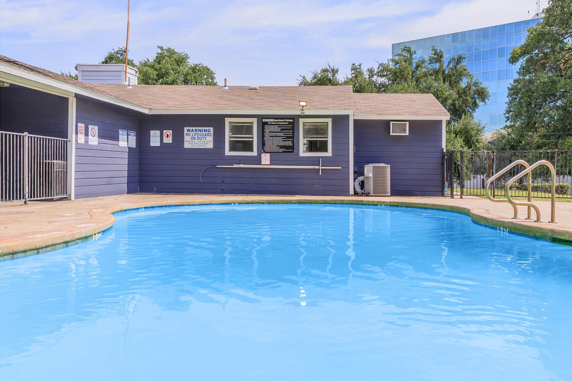 A clear blue swimming pool with a surrounding deck, a safety fence, and a wooden building nearby. The building features windows and signage, indicating pool rules. In the background, a modern glass building can be seen against a blue sky with some clouds.