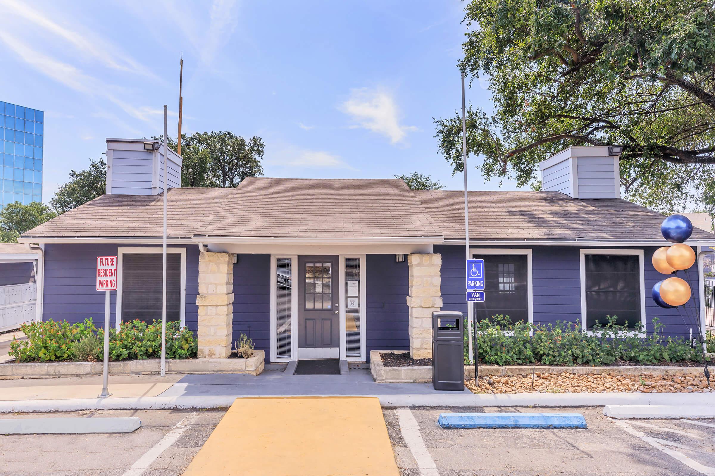 A single-story building with a dark blue exterior and light-colored stone accents. It features a pitched roof, two chimneys, and landscaped greenery in front. There are signs indicating "Public Parking" and "Handicapped Parking." A sidewalk leads to the entrance, with decorative balloons nearby.