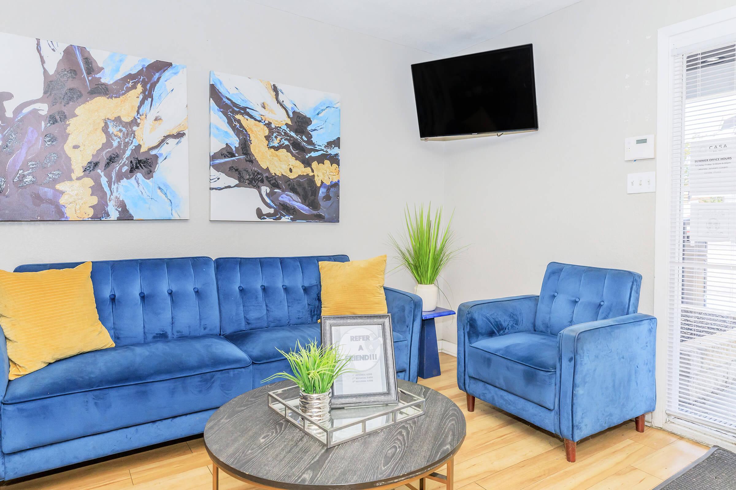 A cozy living room featuring a blue velvet sofa and armchair with yellow accent pillows. Abstract art pieces in blue and gold hang on the wall, and a television is mounted above. A circular coffee table with a green plant centerpiece sits on a warm wooden floor. Natural light streams in through a window with blinds.
