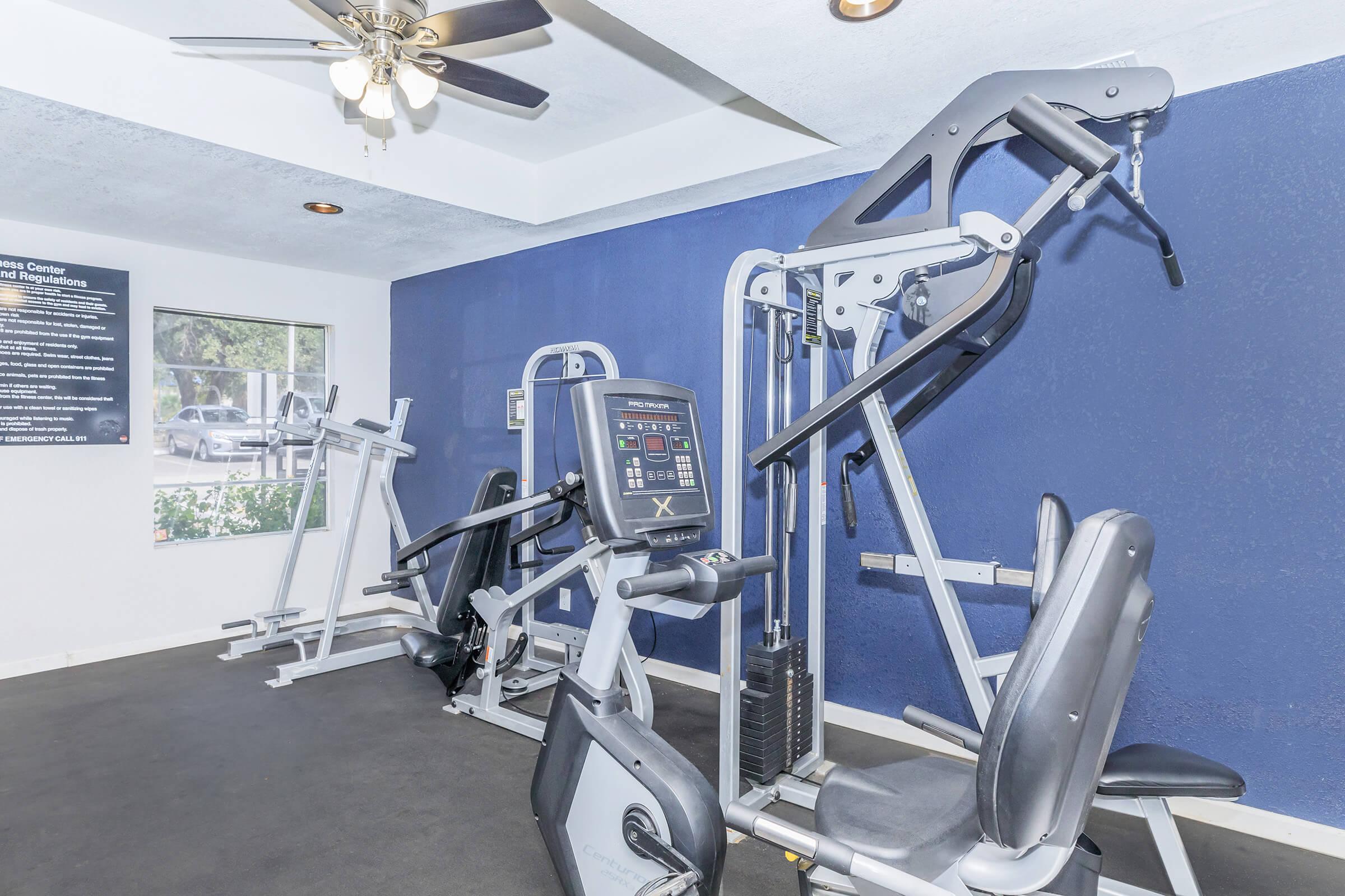 A modern gym space featuring various exercise machines, including a weight machine and an elliptical trainer. The room has a blue accent wall and a ceiling fan, creating a comfortable workout environment. Natural light is coming in through a window, illuminating the fitness area.