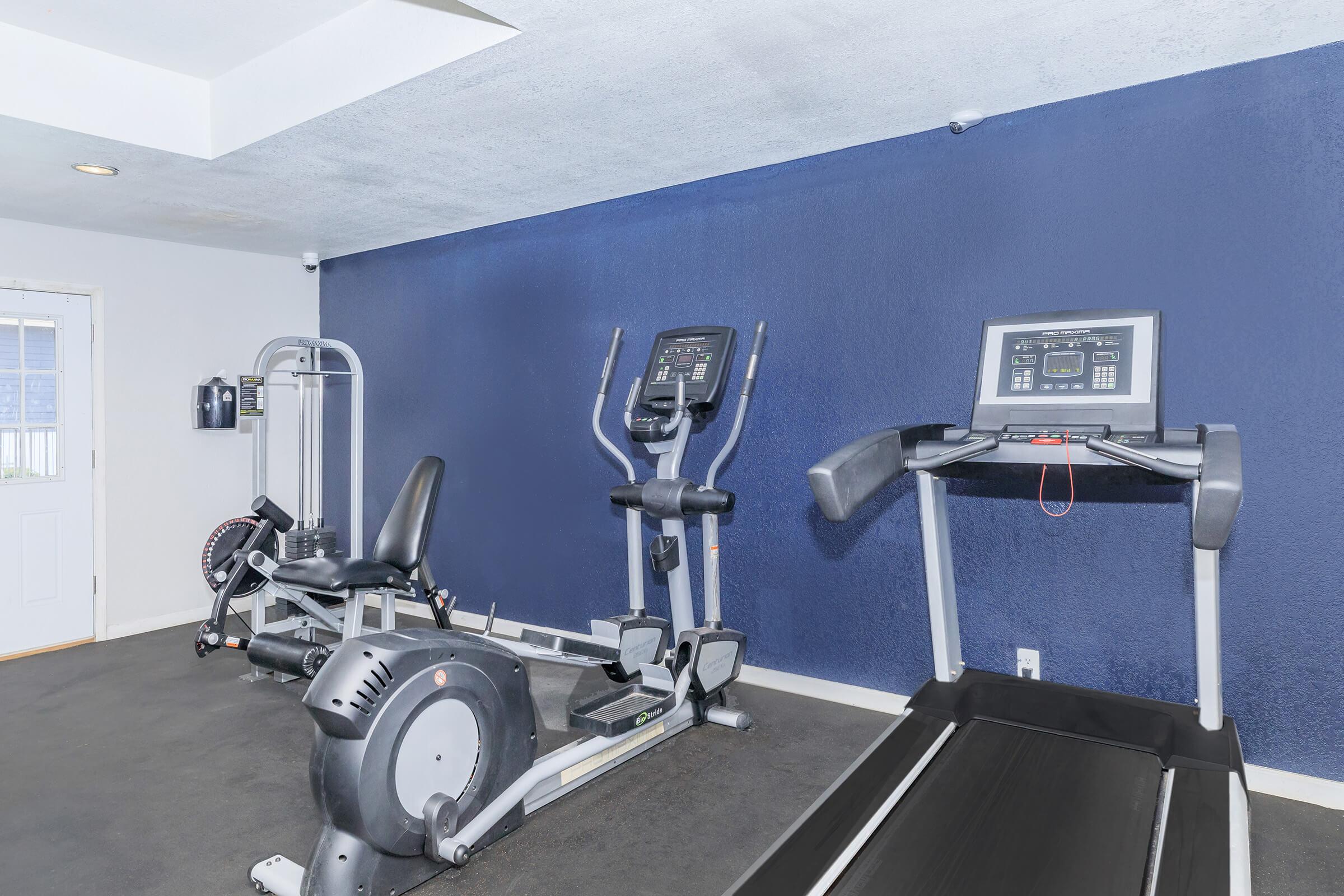 A fitness room featuring a treadmill, an elliptical machine, and a stationary bike against a dark blue wall. The space has a modern design with a rubber floor and ample lighting. A door leads to another area, enhancing the gym's accessibility.