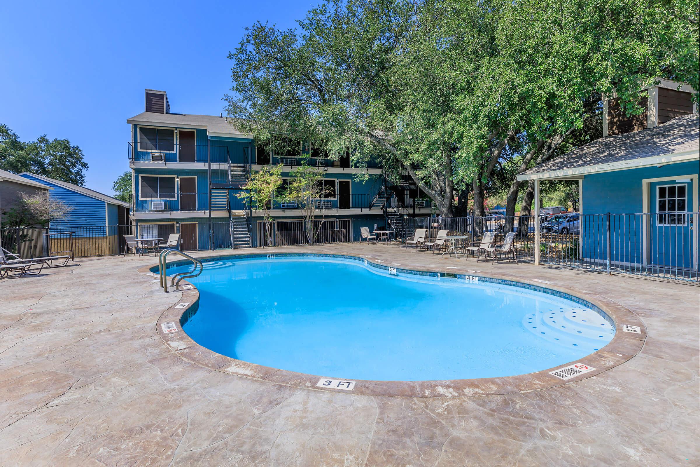 A clear blue swimming pool surrounded by a stone deck, lounge chairs, and shade from nearby trees. In the background, there are two-story buildings with blue siding and balconies. The sky is bright and sunny, creating a refreshing atmosphere.