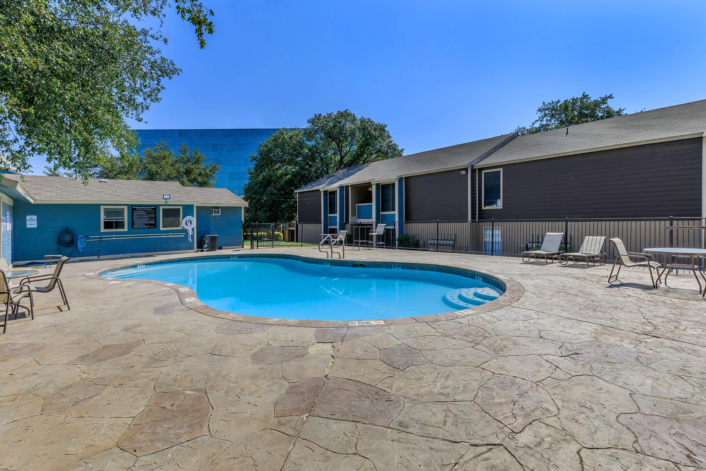 A clear blue swimming pool surrounded by a stone patio and lounge chairs. In the background, there's a two-story building with a mix of siding colors, and trees providing shade. A nearby pool house is visible, along with a clear blue sky overhead.