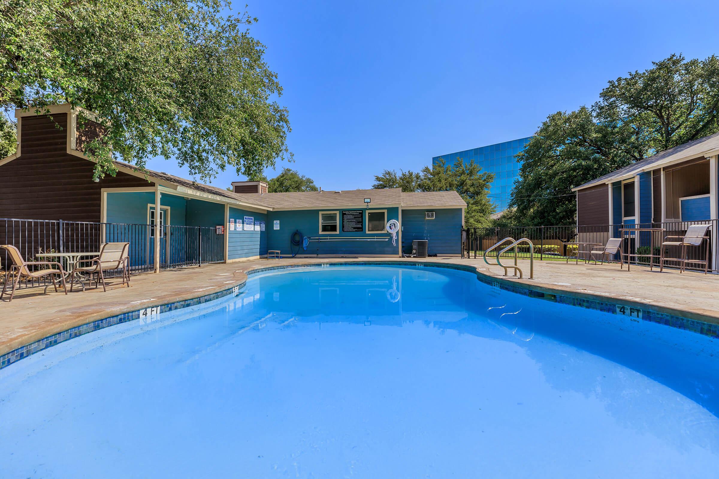 A clear blue swimming pool surrounded by a patio area with lounge chairs, featuring green trees and a nearby building in the background. The scene is bright and sunny, capturing a relaxed outdoor atmosphere.