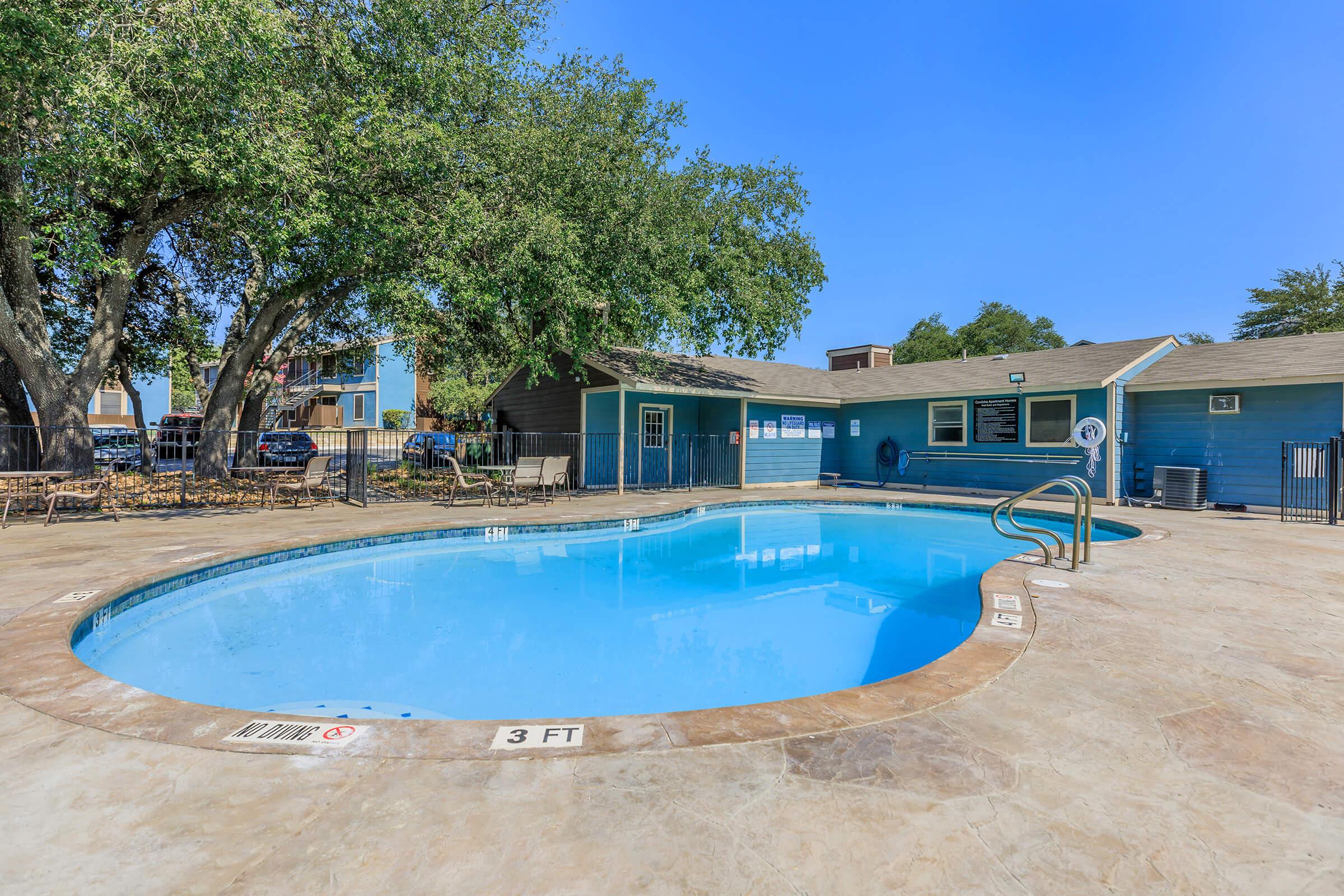 A clear blue swimming pool surrounded by a stone deck and fencing. Lush green trees provide shade nearby. A building with windows is visible in the background, complementing the outdoor setting. The pool area appears inviting and well-maintained, ideal for relaxation and leisure.