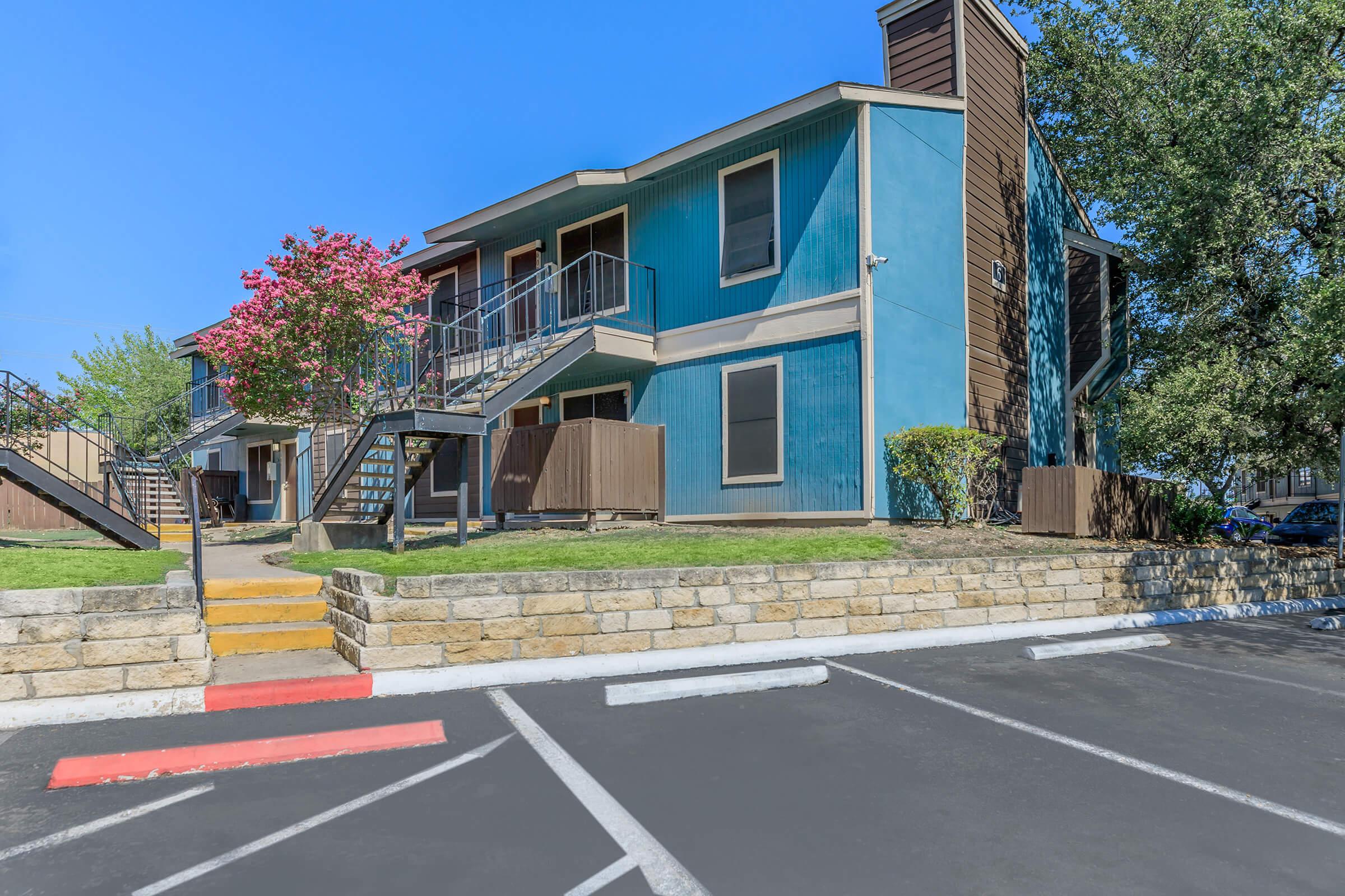 A colorful two-story apartment building with blue and brown exterior, featuring balconies. In front, a landscaped area with a flowering tree and steps leading up to the entrance. A parking lot is visible in the foreground, with marked spaces and a smooth asphalt surface. Clear blue sky overhead.