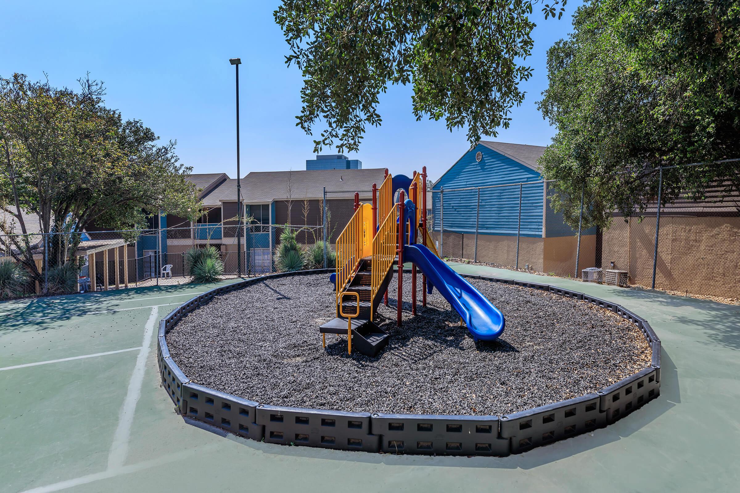 A small playground area featuring a yellow and blue slide, surrounded by rubber mulch and a low black border. The playground is situated on a green court, with trees and a blue building in the background under clear blue skies. Fenced surroundings provide a secure play environment.