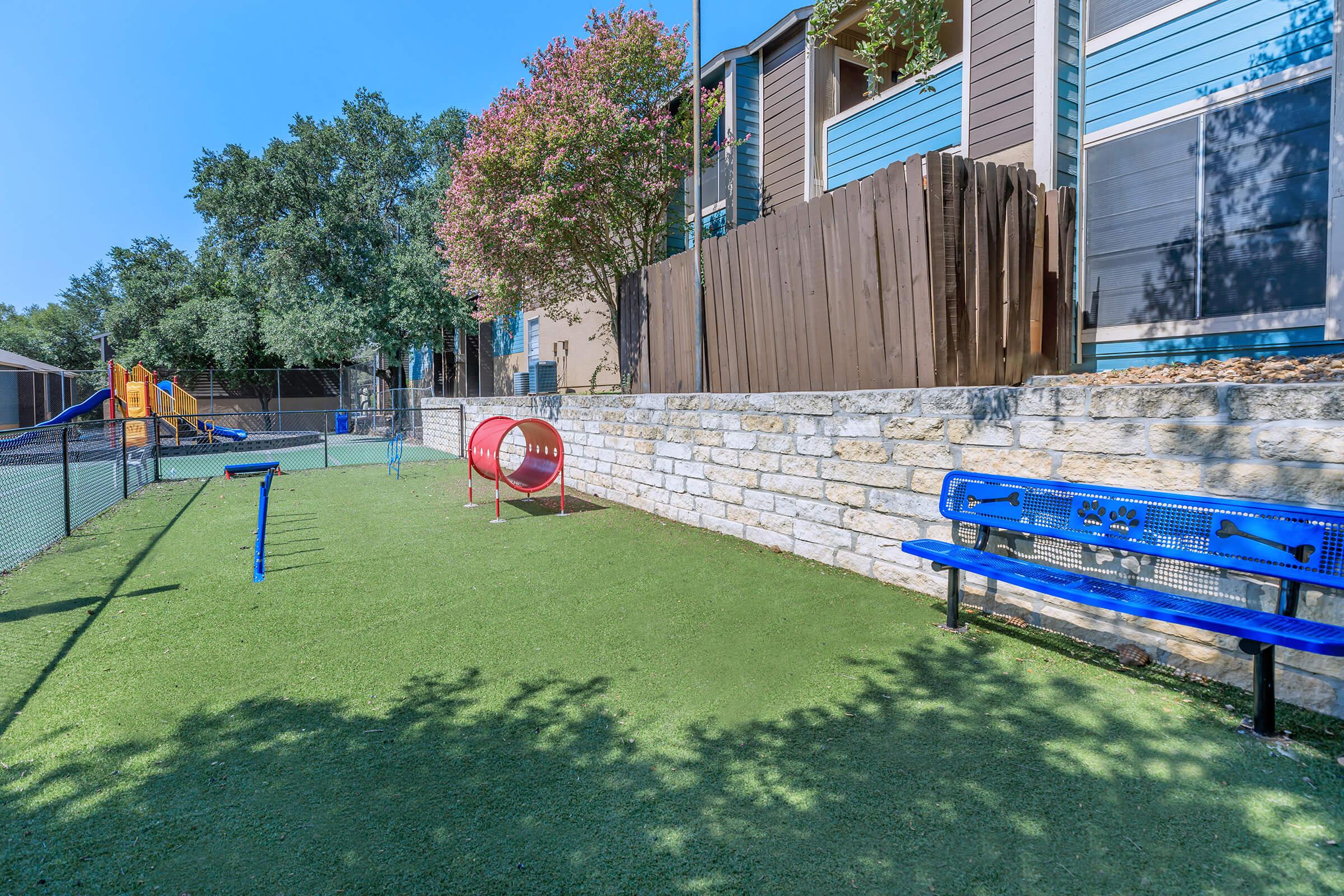 Playground area with artificial grass, featuring a red tunnel-shaped play structure, a blue bench, and a fenced-in space. Background includes a wooden fence and residential buildings with trees providing shade. Clear blue sky above.