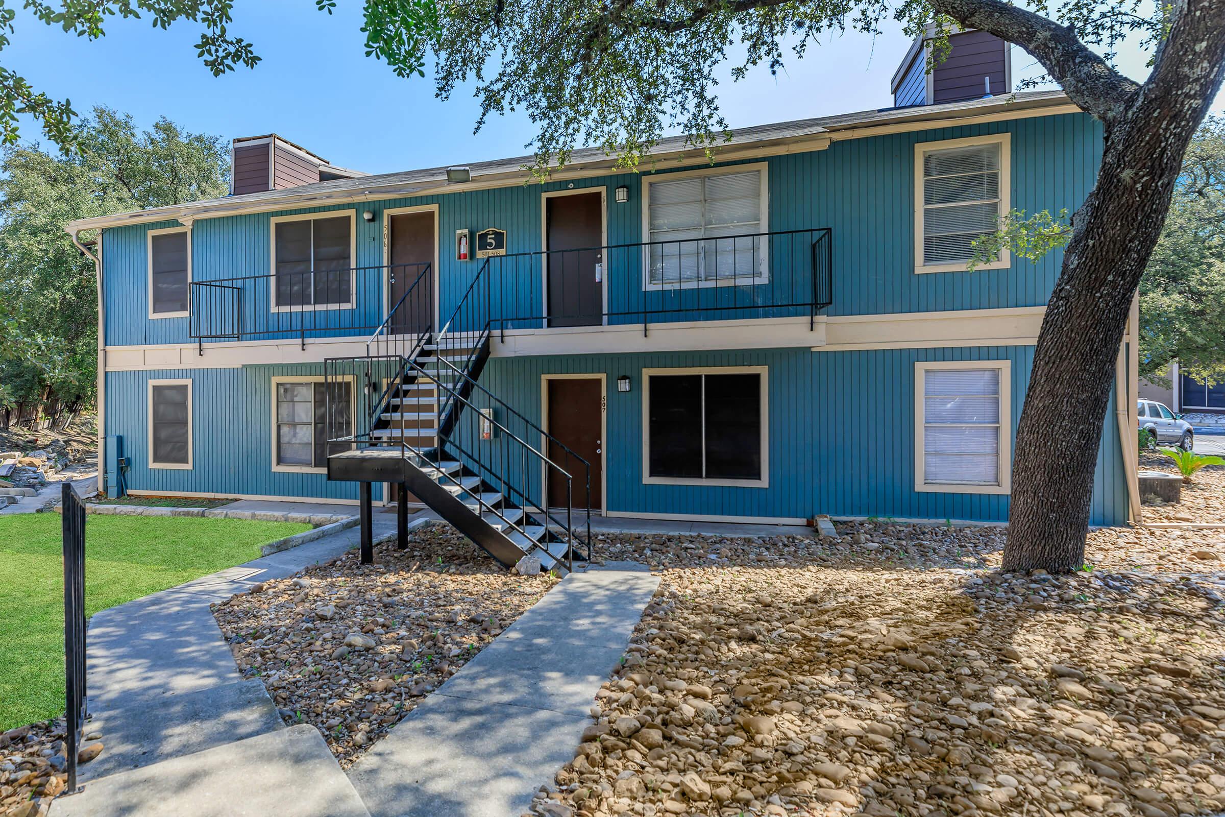 Two-story blue apartment building with multiple entrances and a staircase in front. Surrounding landscaping features grass and decorative stone pathways. A tree is visible to the right, enhancing the outdoor environment.