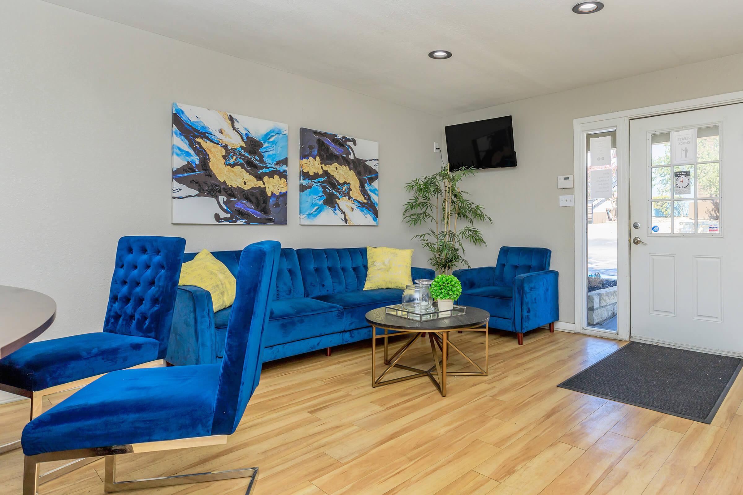 Modern living room featuring a blue velvet sectional sofa with yellow throw pillows, a round coffee table, and vibrant abstract art on the walls. A flat-screen TV is mounted above the entrance door, and there is a potted plant in the corner, creating a bright and inviting atmosphere.