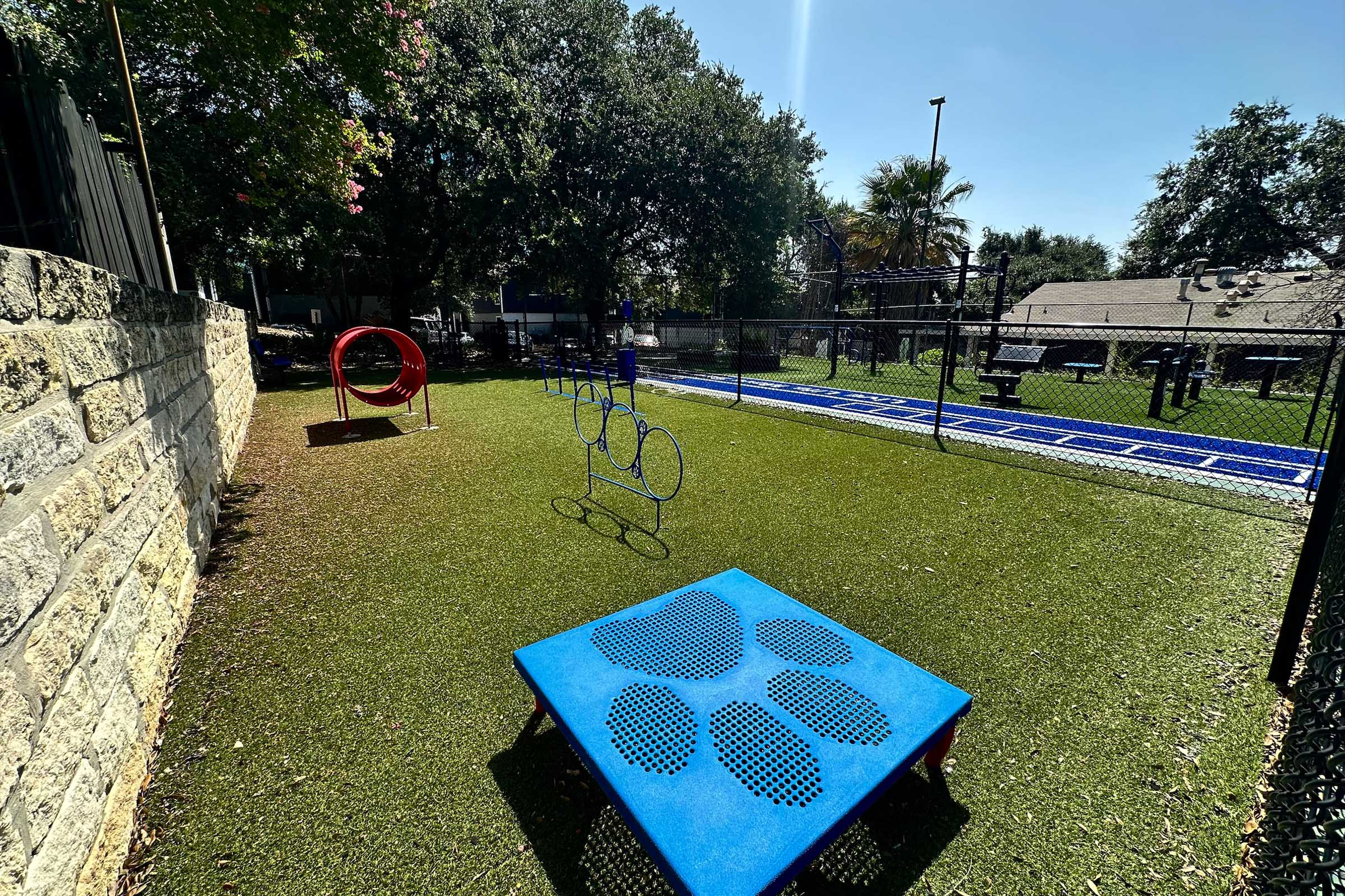 A vibrant, fenced dog play area featuring artificial grass. There are several play structures: a blue platform with paw prints, a red tunnel, and agility hoops, along with a blue running path in the background, surrounded by trees and buildings. The sky is clear and sunny.