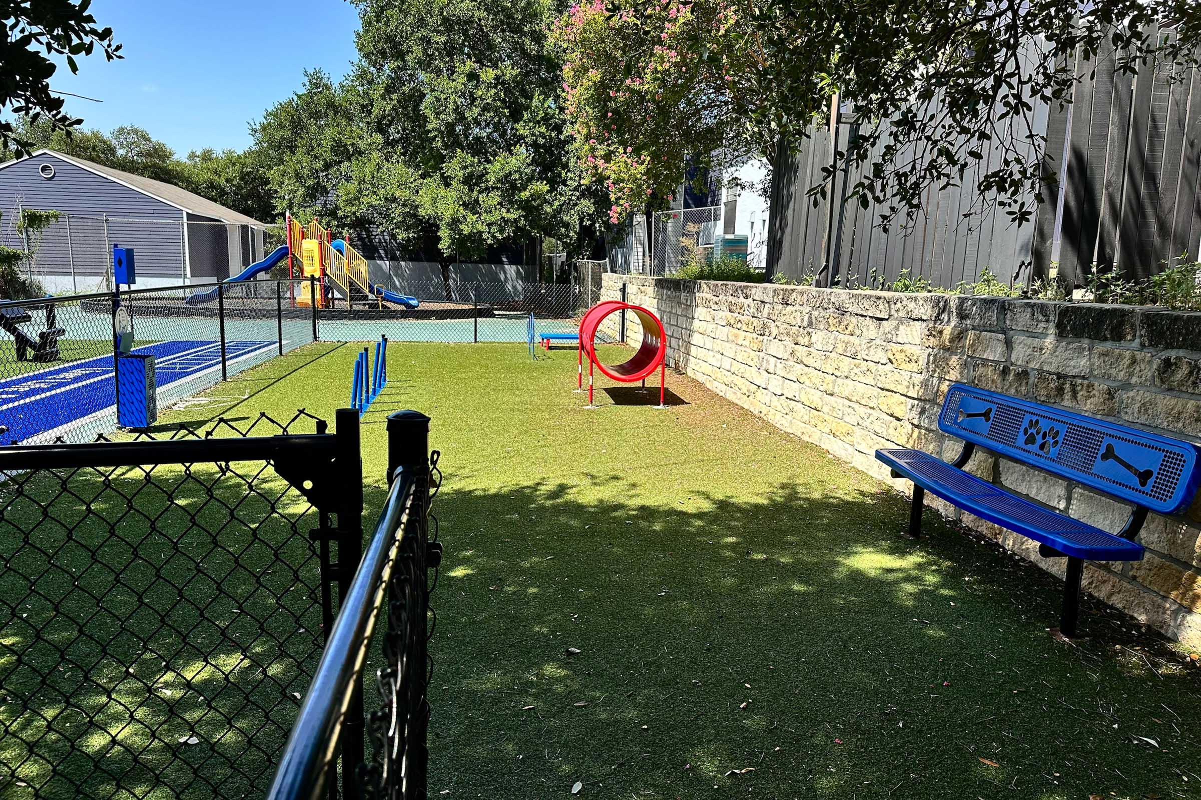 A brightly colored playground area featuring a climbing structure, slide, and various play equipment. There is a fenced grass area with a circular tunnel and a blue bench along the edge. The setting includes trees and a well-maintained landscape in a sunny environment.