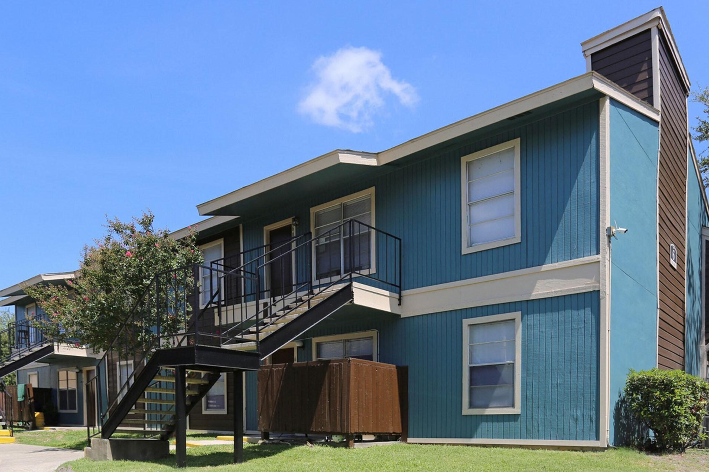 Two-story apartment building with a blue exterior, featuring a staircase on the left. Grass surrounds the building, and a small tree adds greenery. There are paired units visible, each with windows and a wooden stairwell leading to the upper floor. A clear blue sky is in the background.