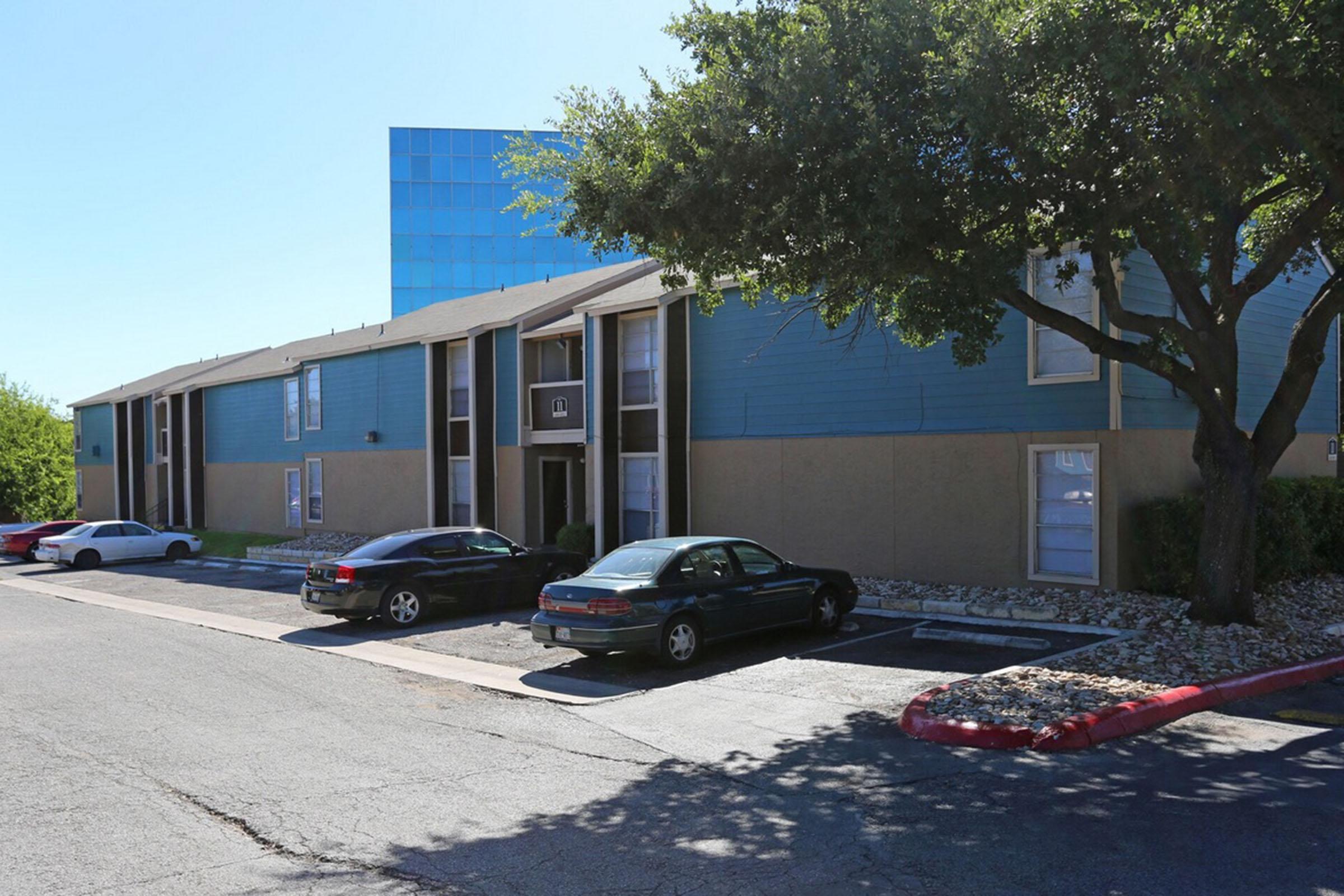 A multi-story apartment building featuring blue and beige siding, surrounded by trees. Two parked cars are visible in front of the building, alongside a gravel area. The background includes a modern glass building, contrasting with the older architectural style of the apartment complex.