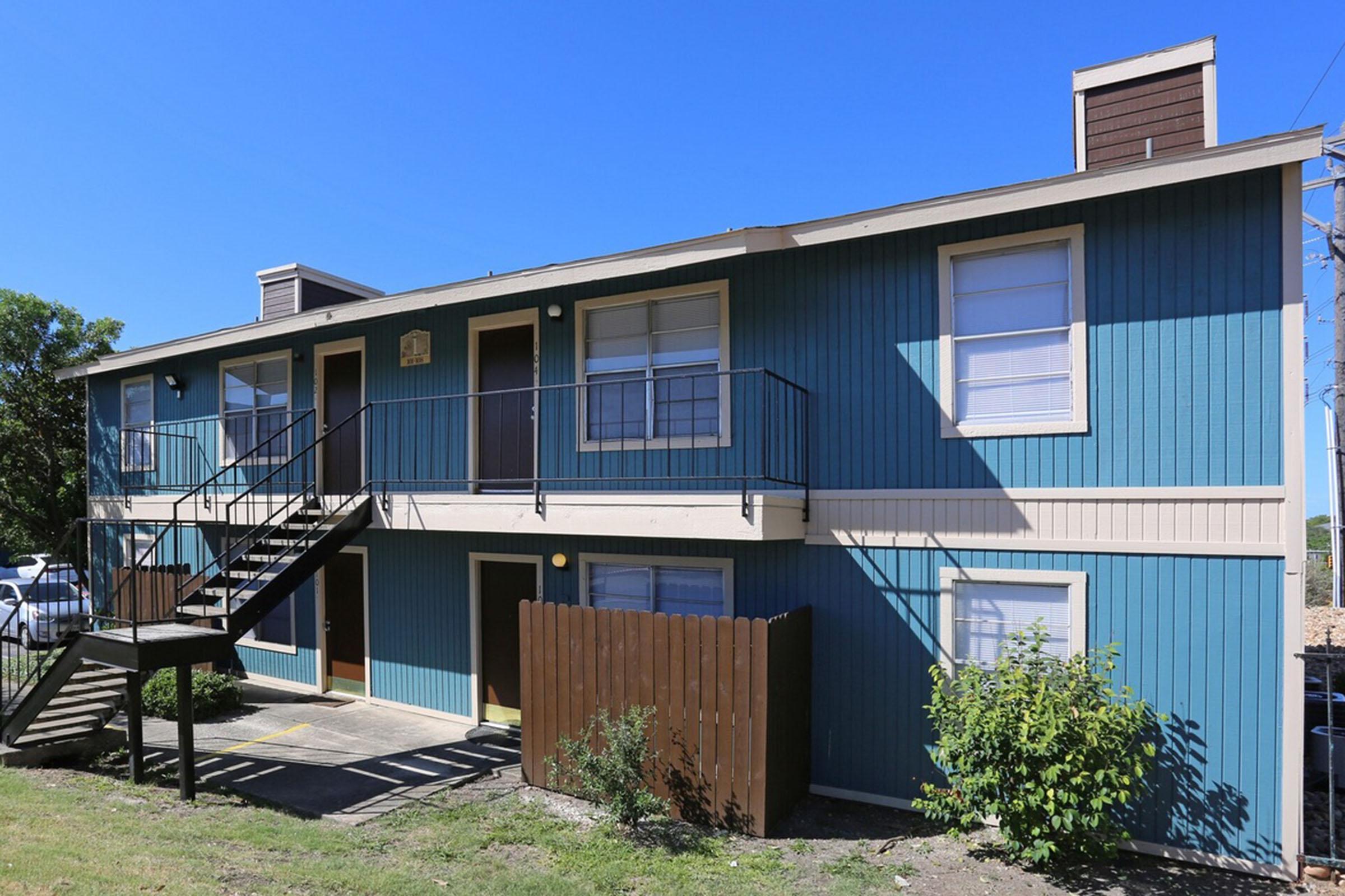 Two-story apartment building with blue and beige exterior. It features a stairway leading to the second floor, multiple windows, and a small grassy area in front. A wooden fence is visible on one side, along with a clear blue sky overhead.